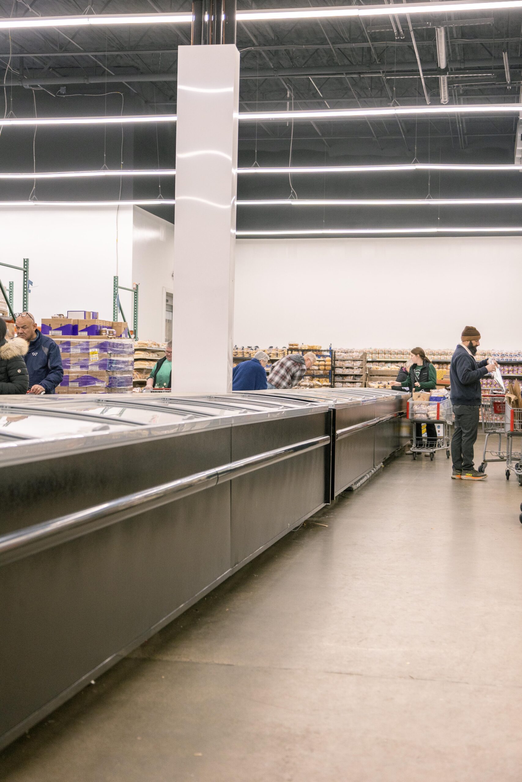 A busy grocery store interior showing shoppers browsing frozen food sections and bakery items, with modern lighting and organized shelves in the background.