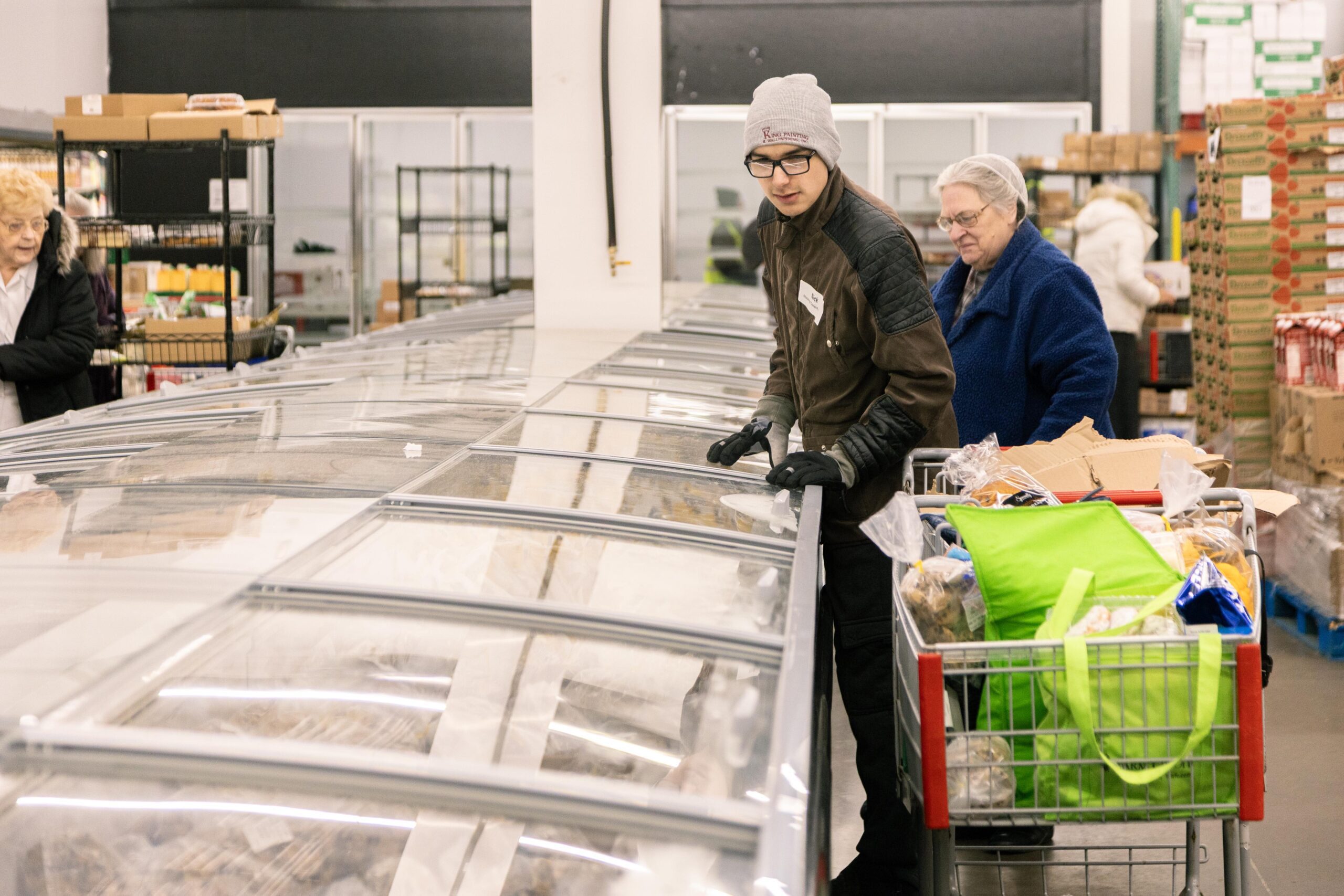 Young man in a brown jacket and gloves examining food items in a grocery store freezer while an older woman in a blue coat looks on, with shopping carts filled with groceries in the foreground.