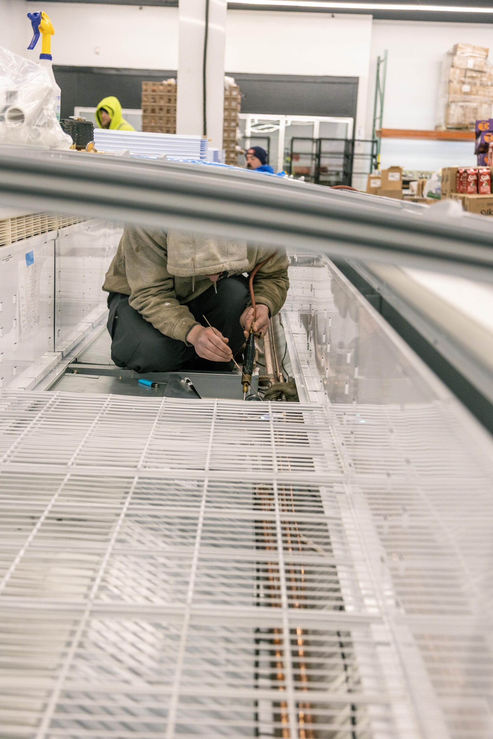 Technician performing maintenance inside a commercial freezer, using tools to work on refrigeration components, with another worker visible in the background.