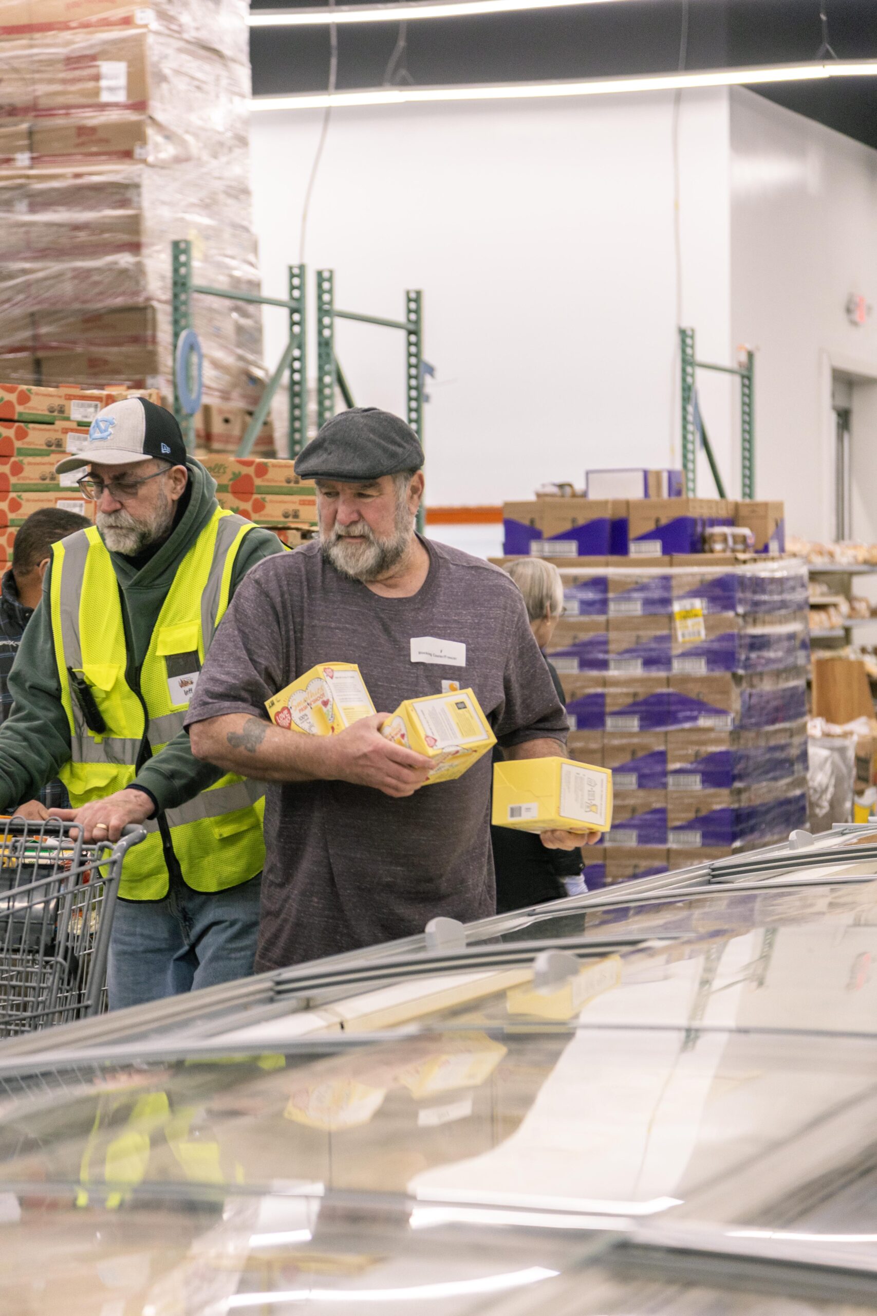 Two men shopping in a grocery store, one wearing a yellow safety vest pushing a cart while the other carries several yellow food boxes. The background features shelves stocked with various products, creating a bustling shopping environment.