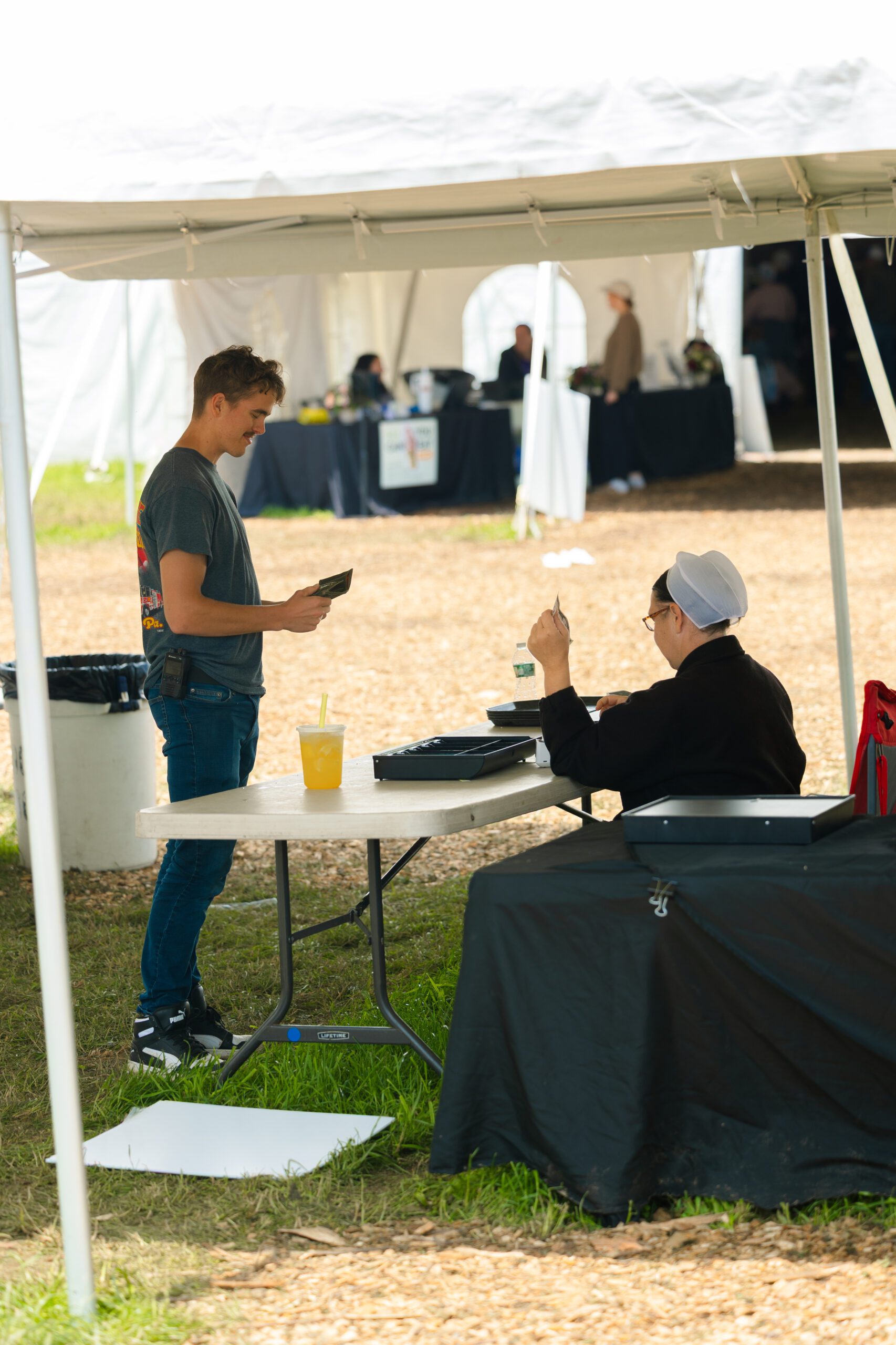 A young man making a payment at a vendor table while an older woman in a traditional outfit assists him at an outdoor market event.