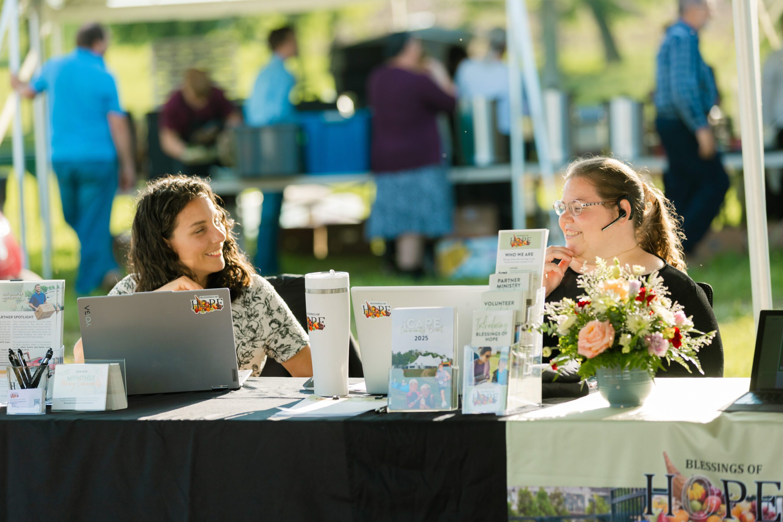 Two women engage at a volunteer information booth under a tent, surrounded by promotional materials for Blessings of Hope. One woman is using a laptop while the other wears a headset and smiles. A floral arrangement adds a welcoming touch to the booth, which includes brochures about volunteer opportunities and community initiatives.