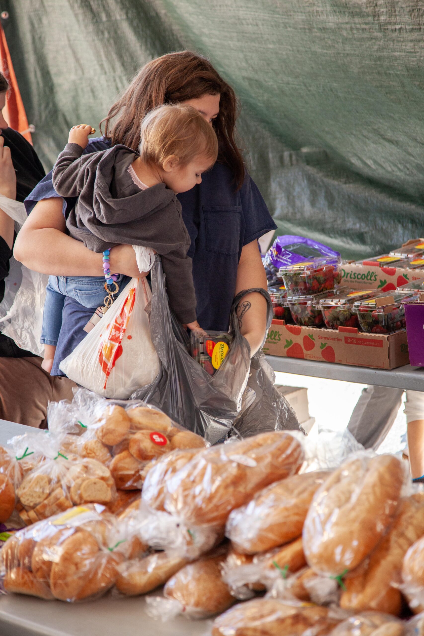 Woman carrying a child while shopping at a market, holding bags of groceries including fresh bread and strawberries, with various baked goods displayed on a table.