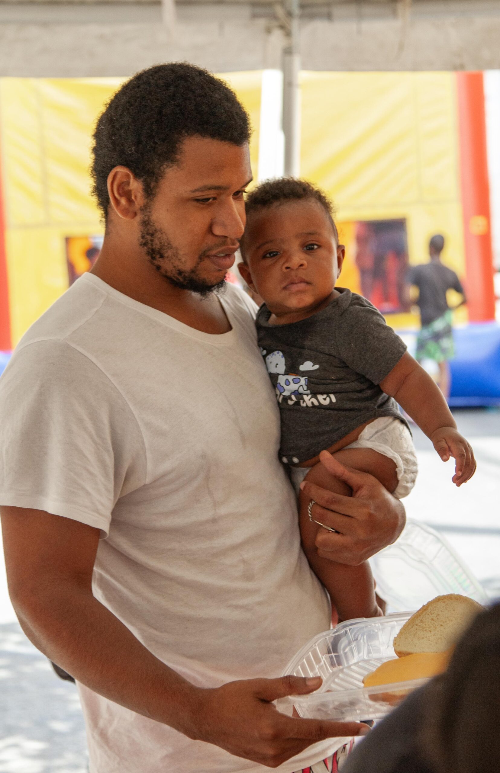 A father holding a baby while carrying a tray with sandwiches at an outdoor event, set against a colorful background of a bounce house.