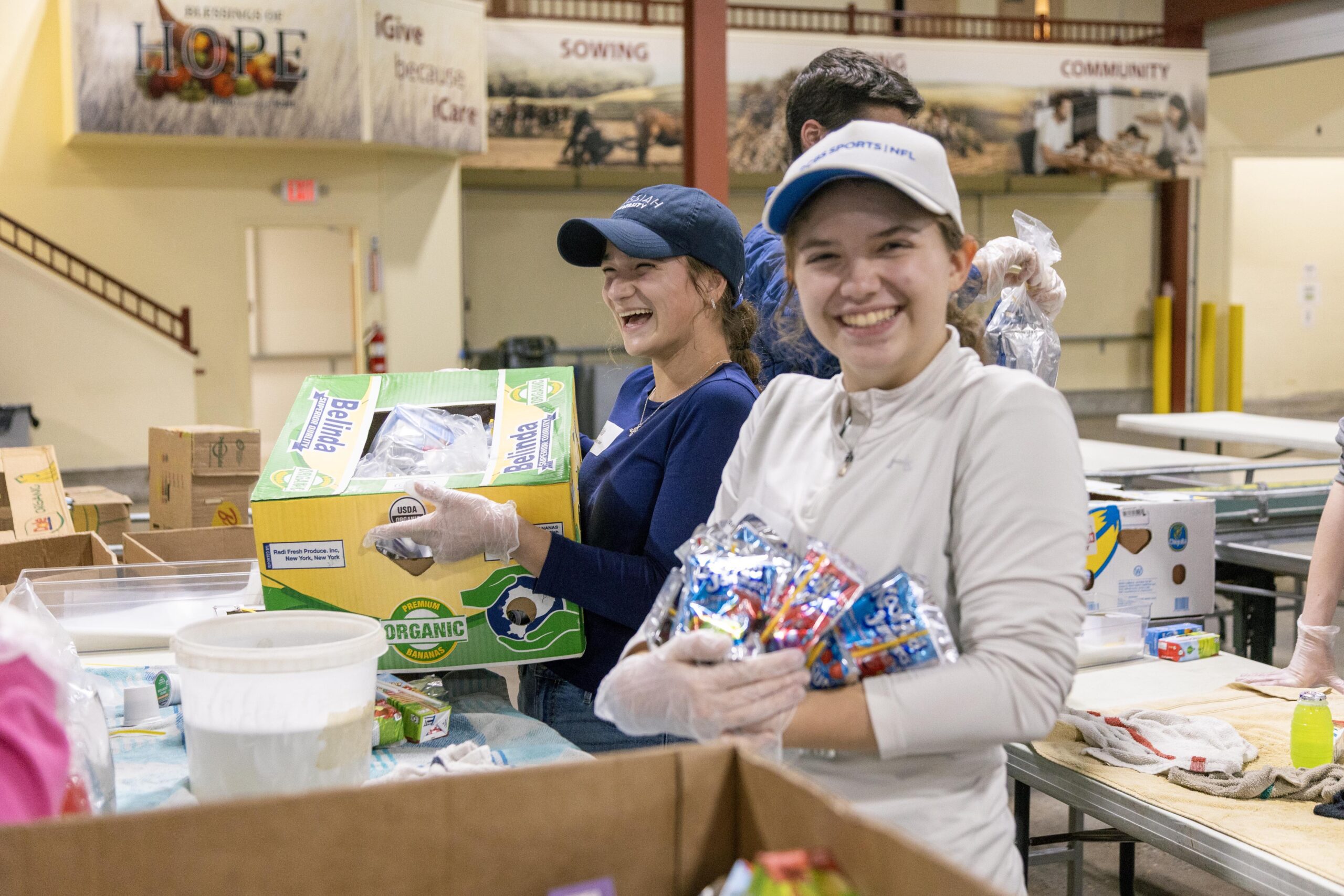 Volunteers at a community food pantry smile while sorting and packing food items, including snacks and organic produce, emphasizing teamwork and generosity in a supportive environment.