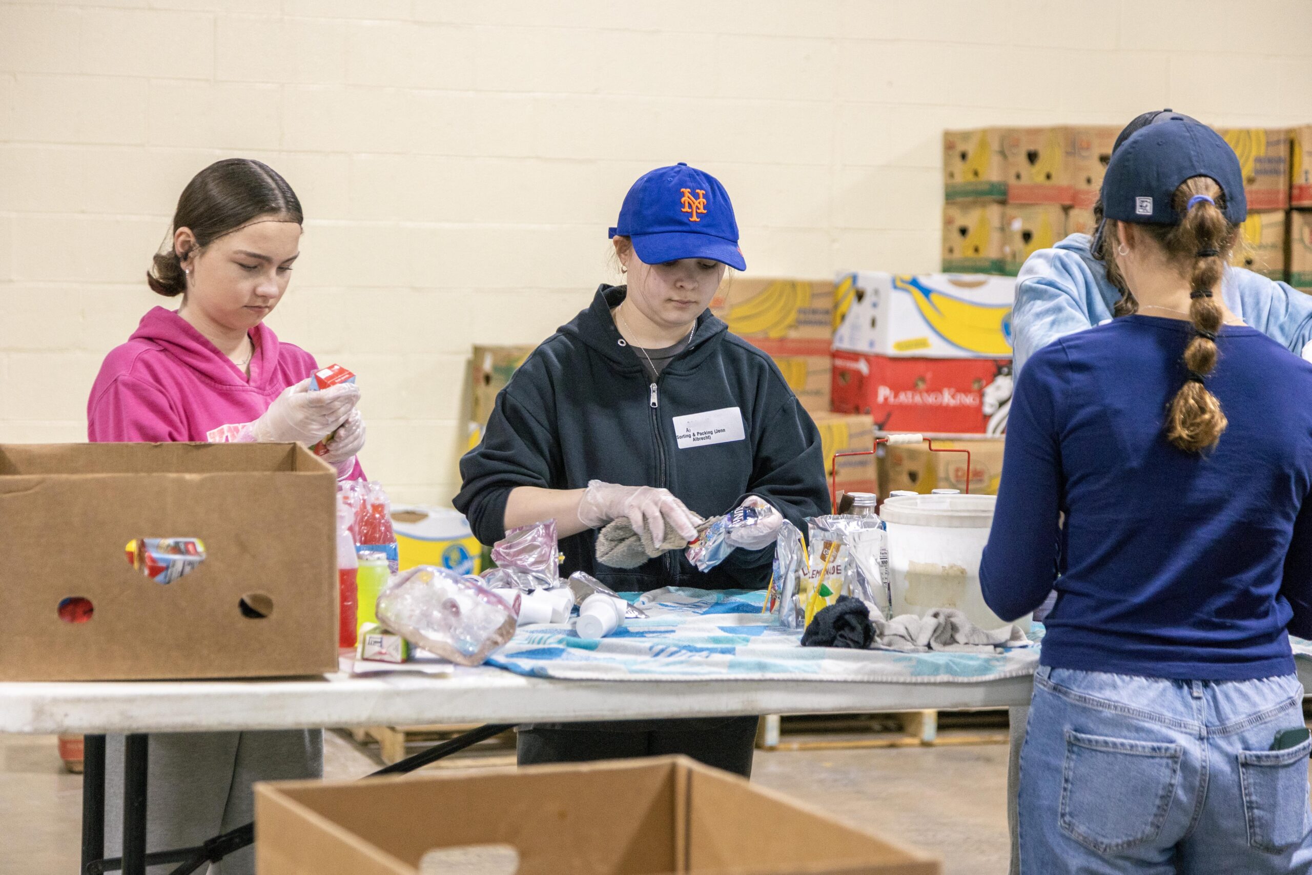 Three young volunteers sorting food items at a community food bank, with cardboard boxes and various food packages visible on a table in a warehouse setting.