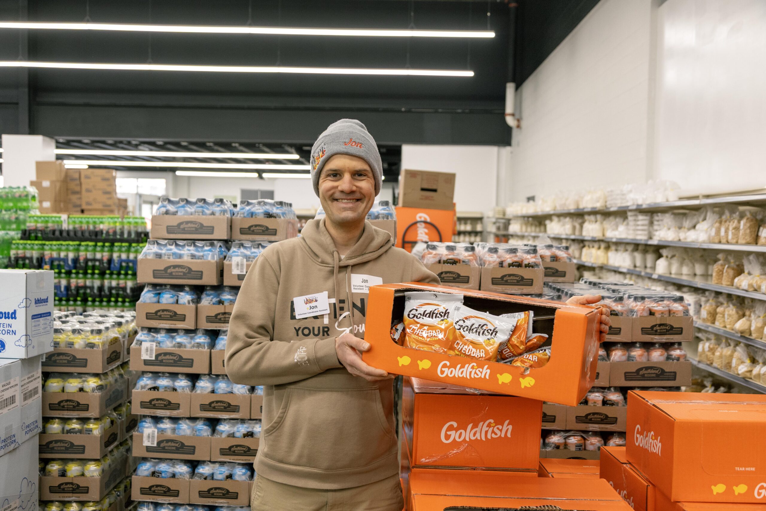 Employee holding a box of Goldfish snacks in a grocery store aisle, surrounded by stacks of soda cans and packaged goods.