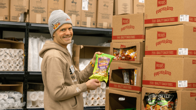 Smiling man in a gray beanie holding a bag of Herr's Sweet Corn Popcorn in a warehouse filled with snack food boxes and supplies.