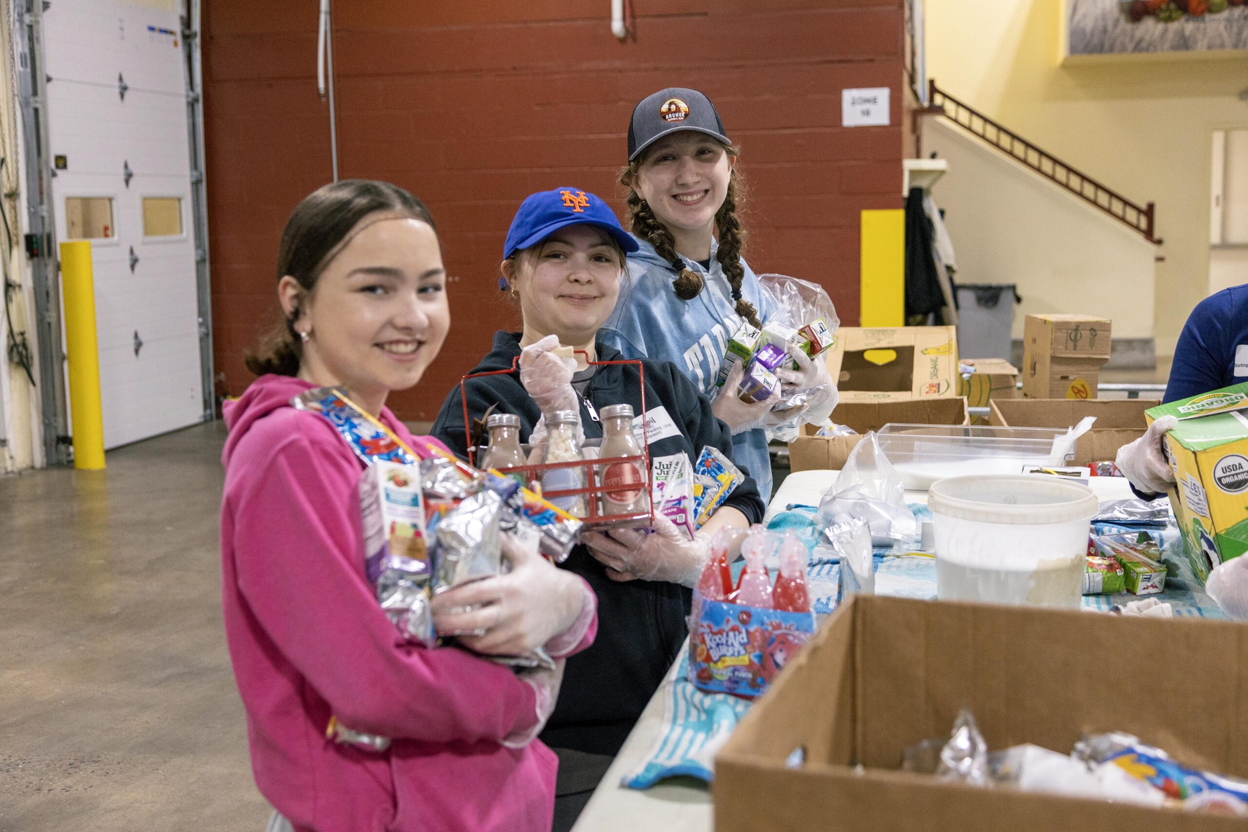 Three young volunteers smiling while holding food items and drinks at a community food distribution center, surrounded by boxes and supplies on a table.