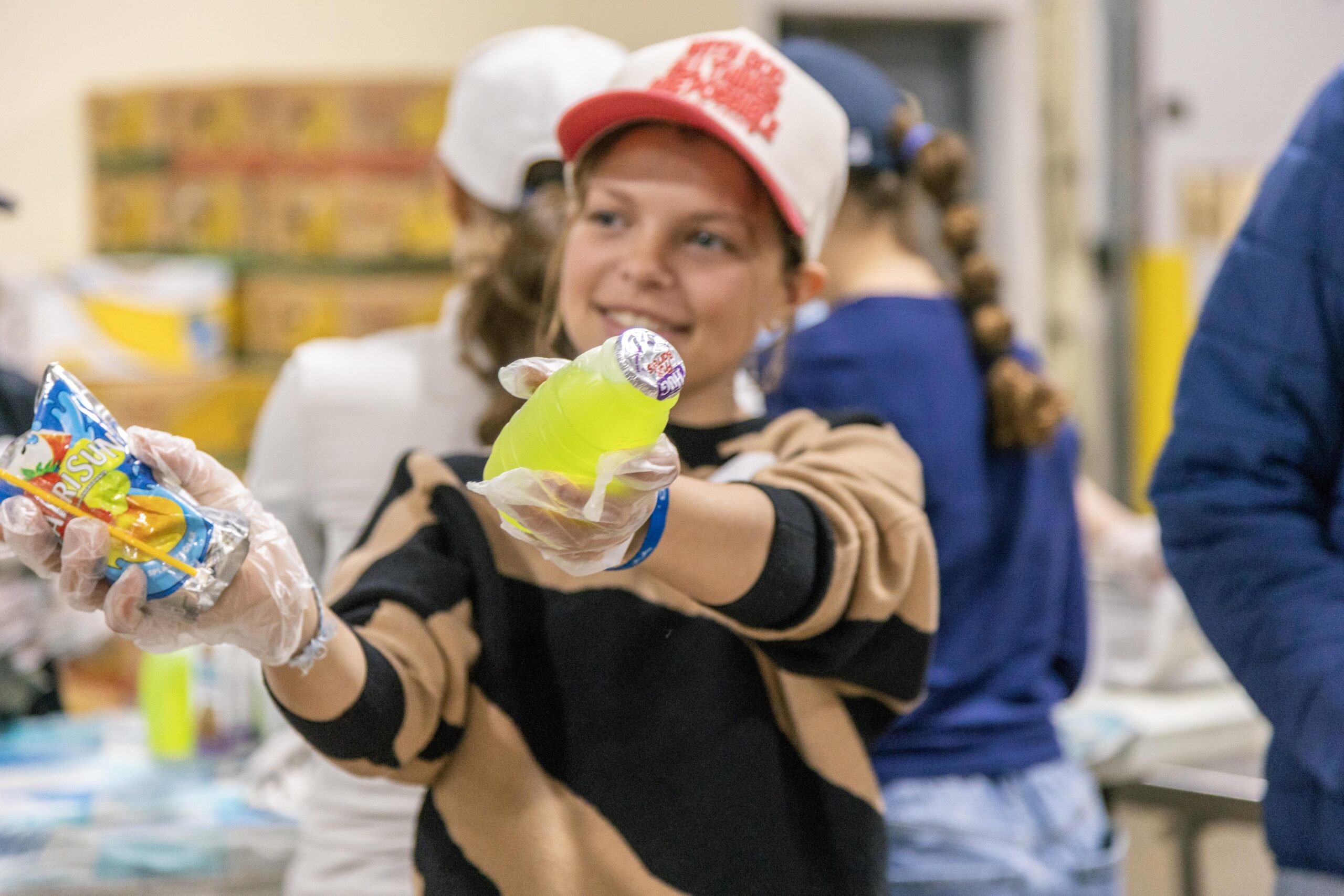 Young girl in a black and tan sweater and a cap, smiling while holding a bottle of bright green drink and a fruit snack, participating in a community food distribution event.