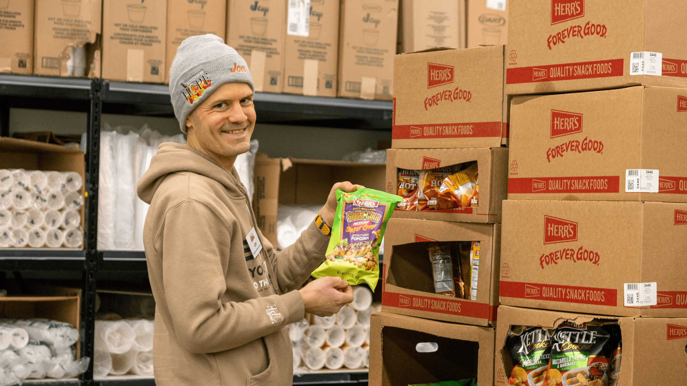 Smiling man in a gray beanie holding a bag of Herr's Sweet Corn Popcorn in a warehouse filled with snack food boxes and supplies.