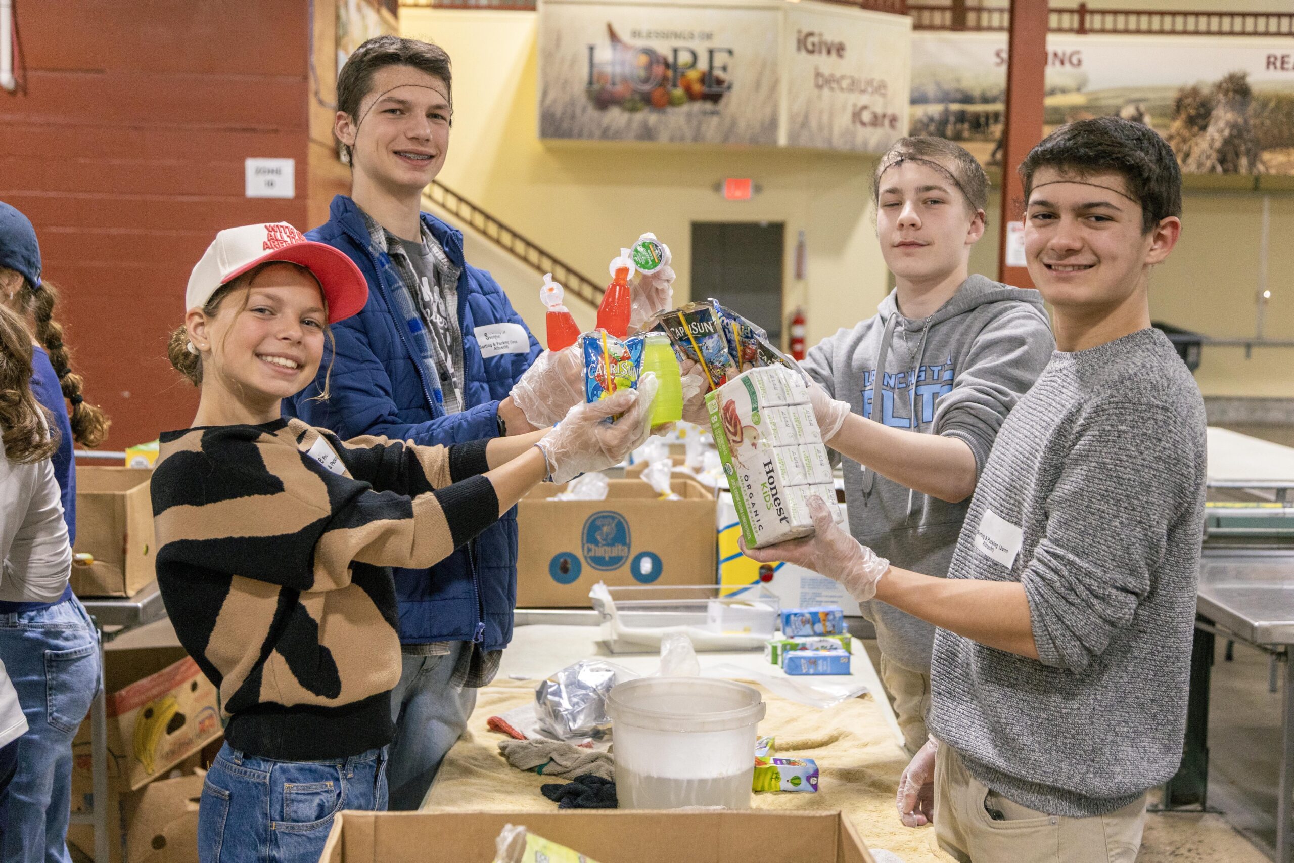 Teenagers volunteering at a food distribution event, smiling as they hold packaged food items, including drinks and snacks, while working together in a community service setting.