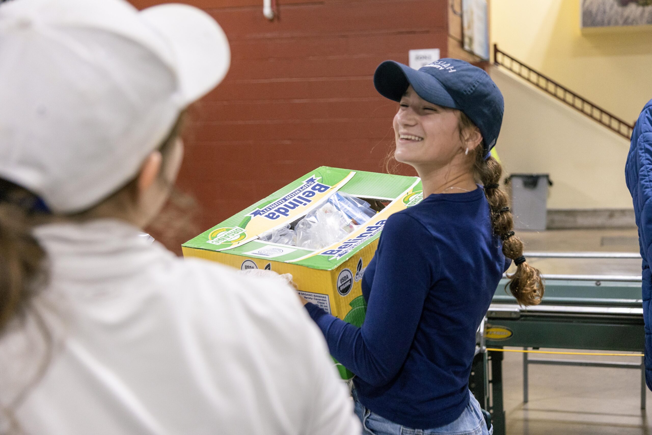 Young woman smiling while carrying a box of Bellita brand products in a warehouse setting, with another person in a white cap visible in the background. The image captures a moment of teamwork and positivity in a community service or food distribution event.