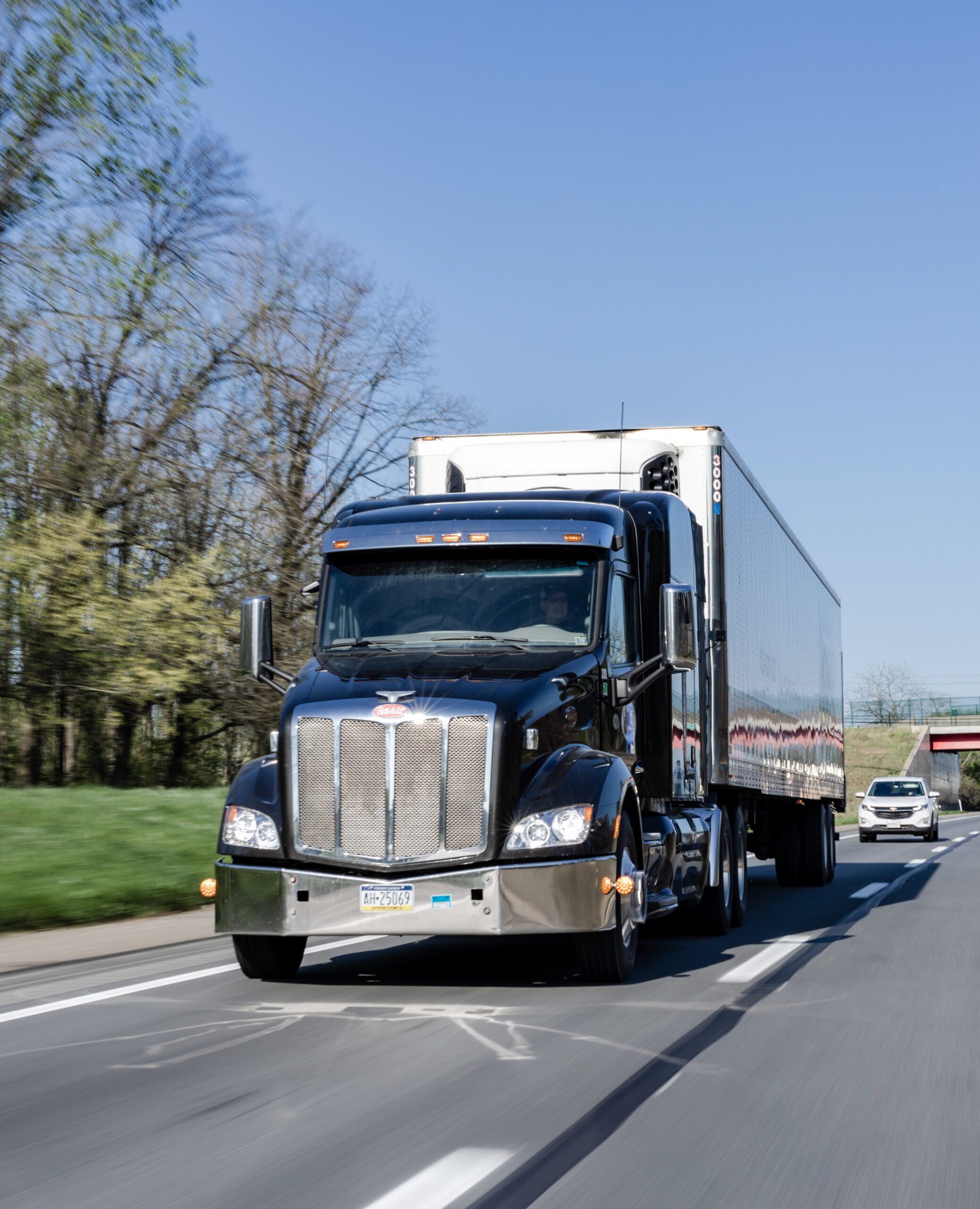 Black semi-truck with a silver trailer driving on a highway, surrounded by greenery and clear blue skies. The truck features a prominent grille and is in motion, showcasing its powerful design.