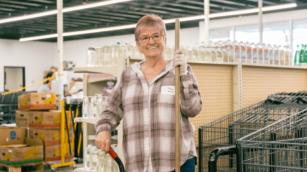 Smiling senior woman in a plaid shirt holding a broom in a store aisle, surrounded by shelves with cleaning products and grocery carts, showcasing community engagement and volunteer work.