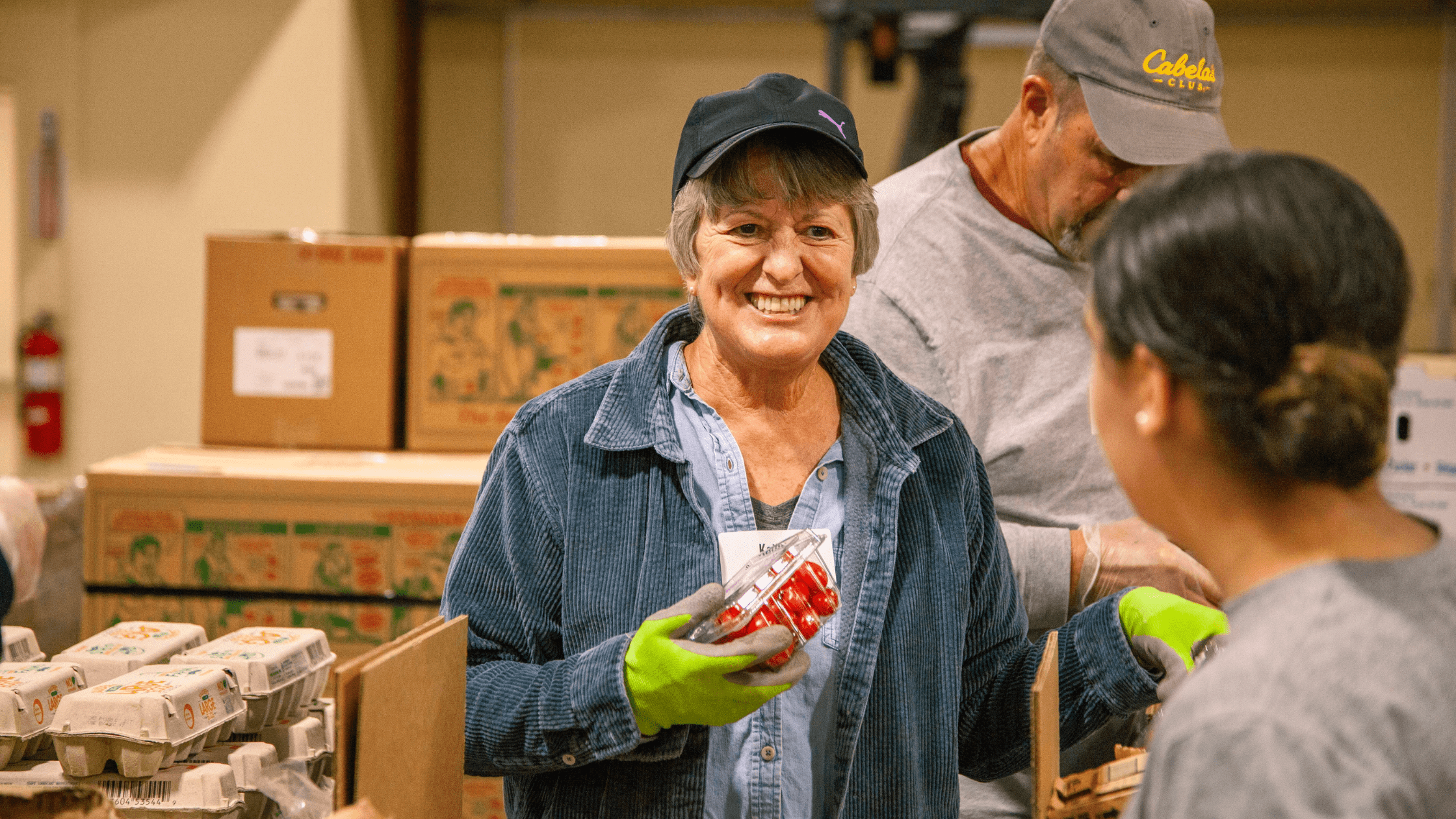 Smiling woman in a blue shirt and gloves holding a container of cherry tomatoes while volunteering in a food distribution center, with boxes of eggs and other volunteers in the background.