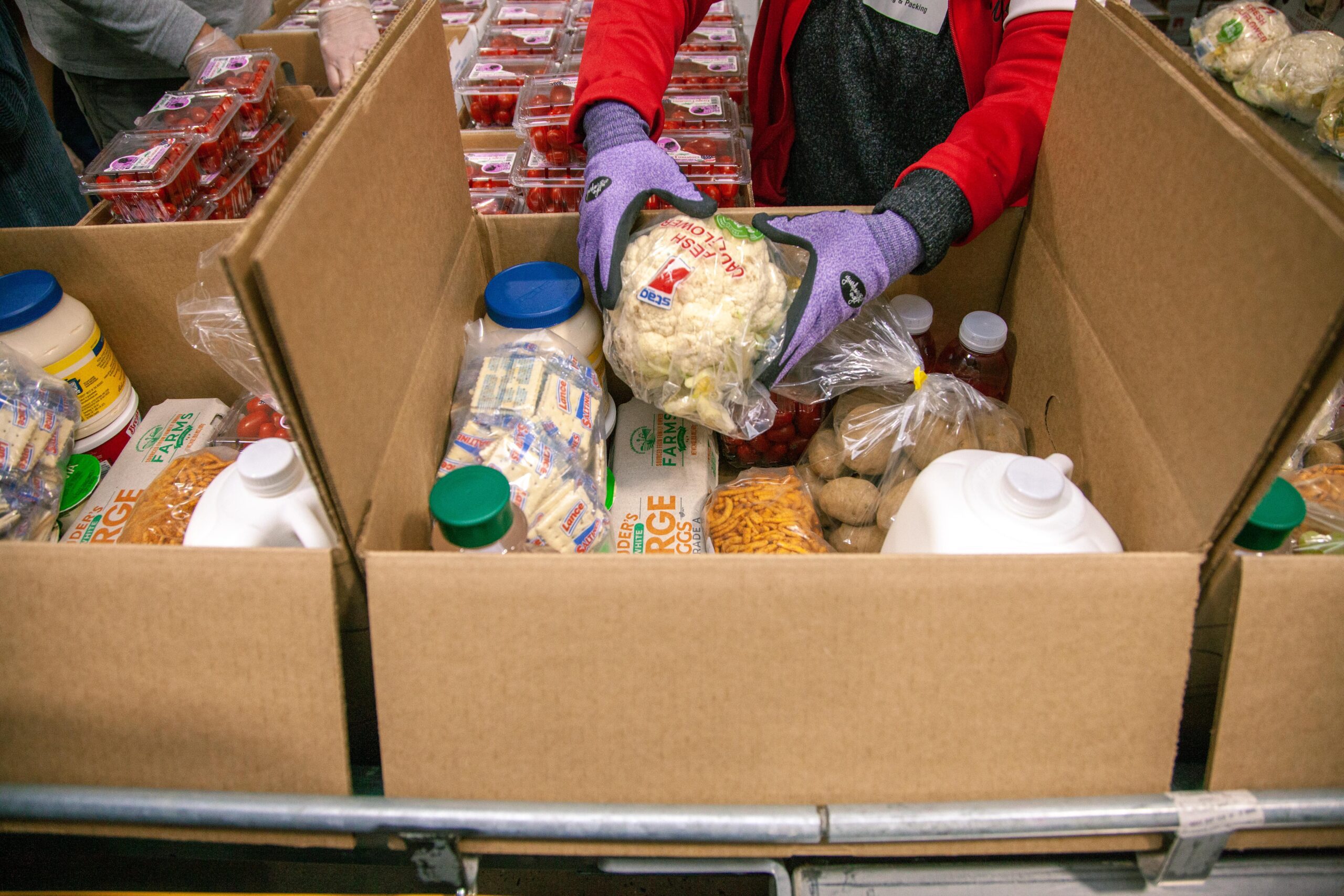 Volunteers packing food items into cardboard boxes, including fresh vegetables, dairy products, and snacks, at a community food distribution event.