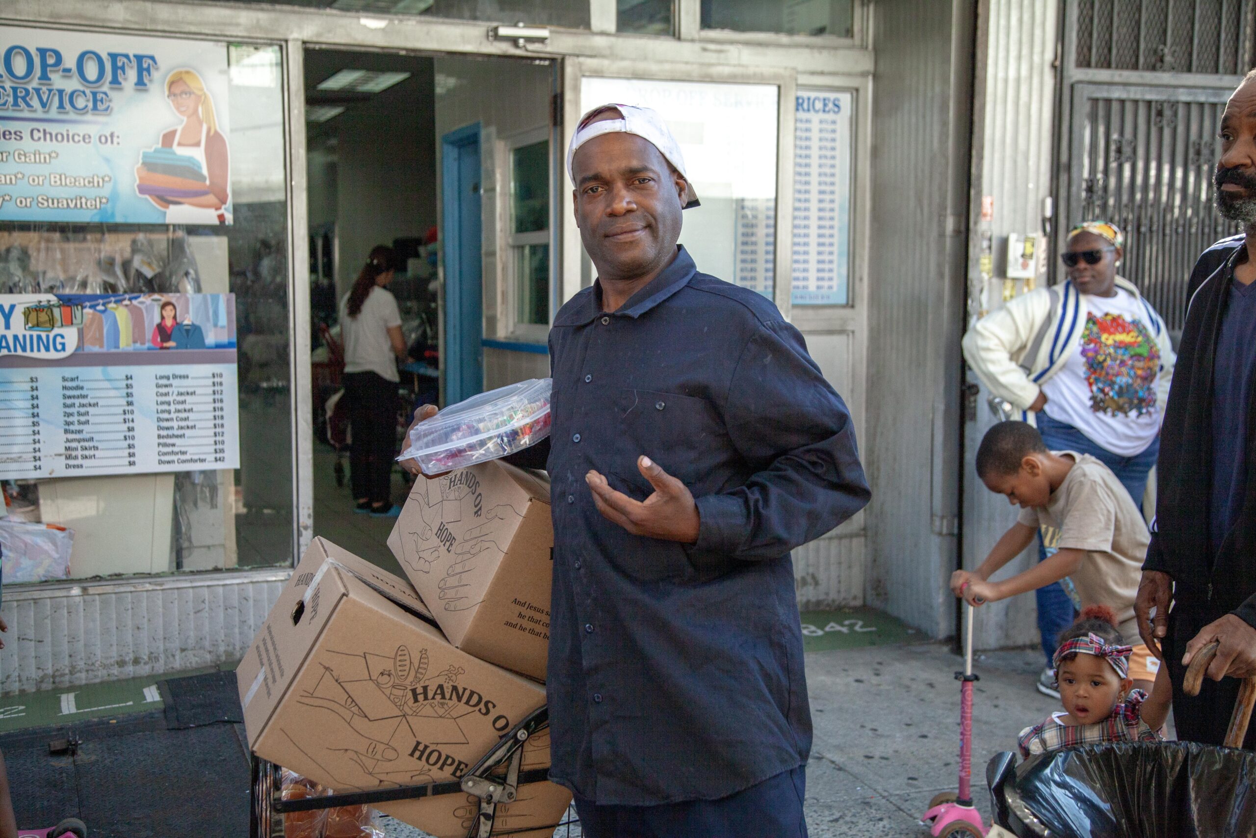 Man standing outside a laundromat holding a container and boxes, with a busy street scene in the background featuring children and adults. The storefront displays laundry service options and pricing information.