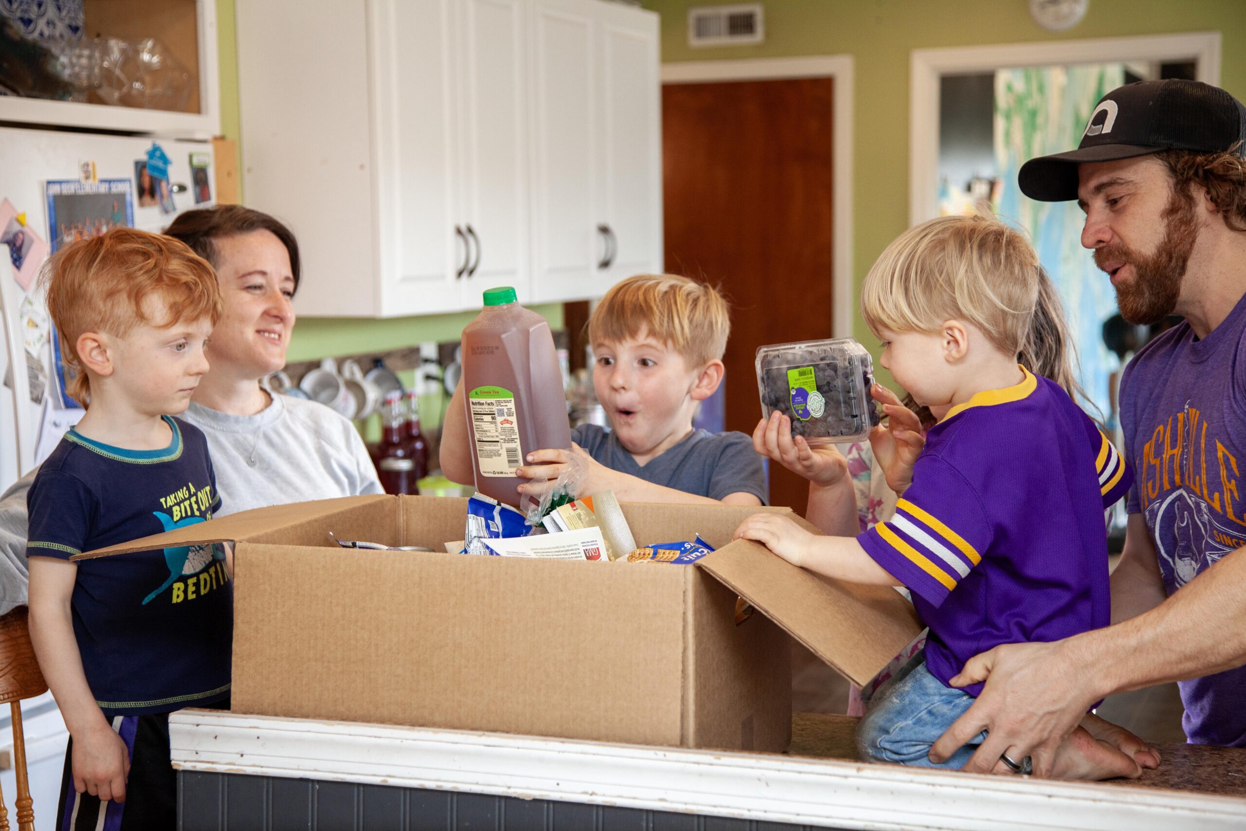 Family unpacking a large cardboard box filled with groceries, including juice and fresh blueberries, in a bright kitchen. Two children express excitement while their parents assist them.