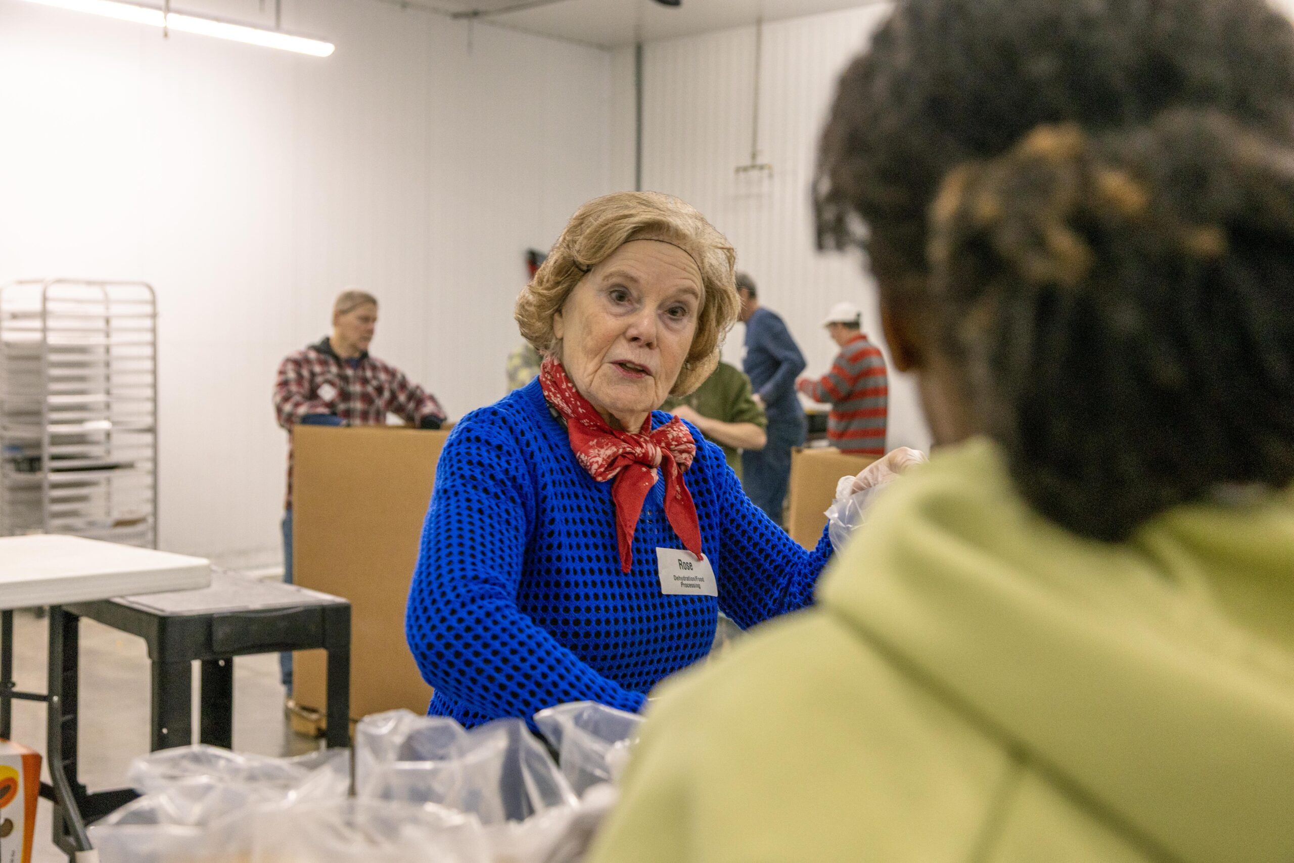 A woman in a blue sweater and red scarf engages in conversation while volunteering at a food distribution center, surrounded by other volunteers. The setting includes tables and boxes, emphasizing community service and teamwork.