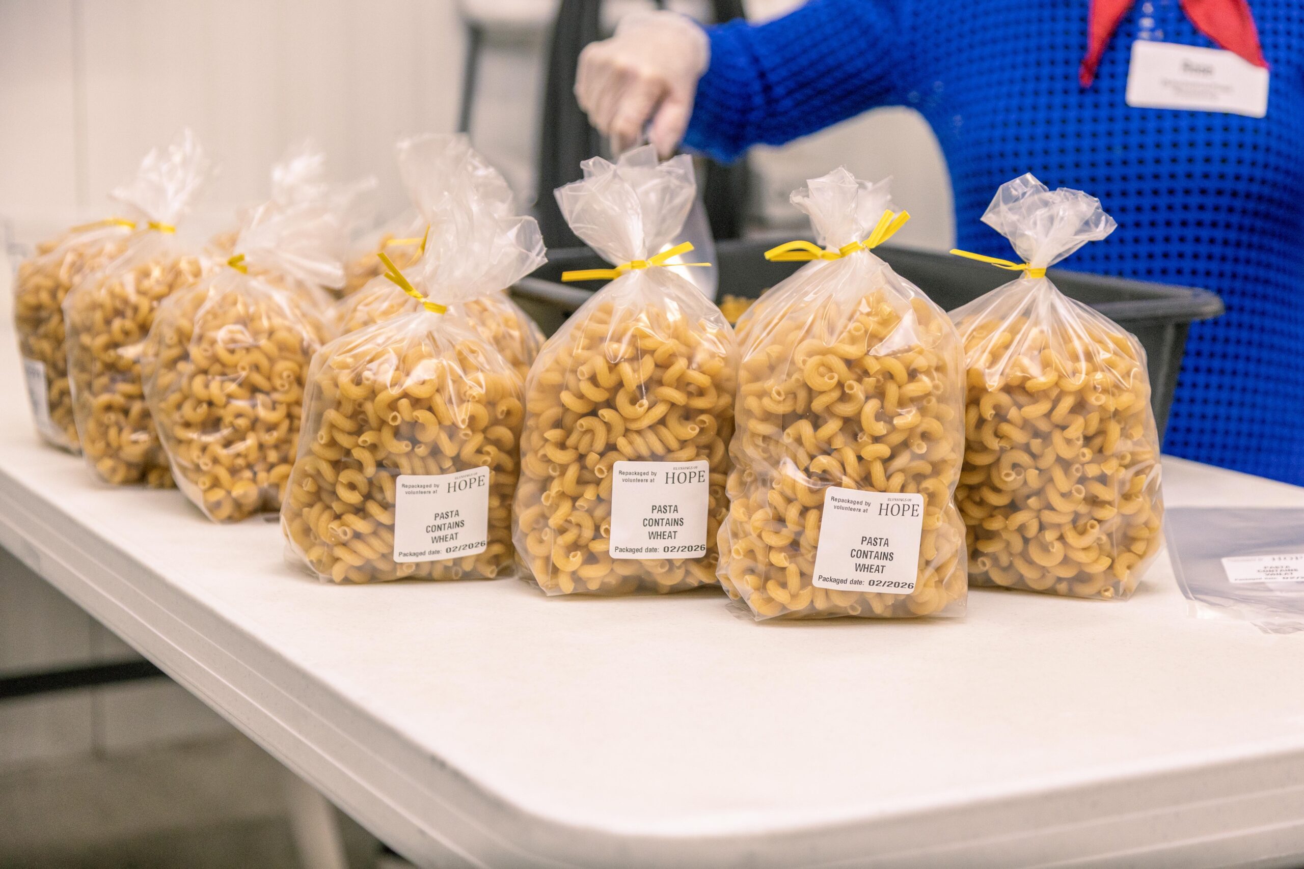 Bags of macaroni pasta packaged for distribution, featuring labels that indicate the product contains wheat, displayed on a table in a community food program setting.