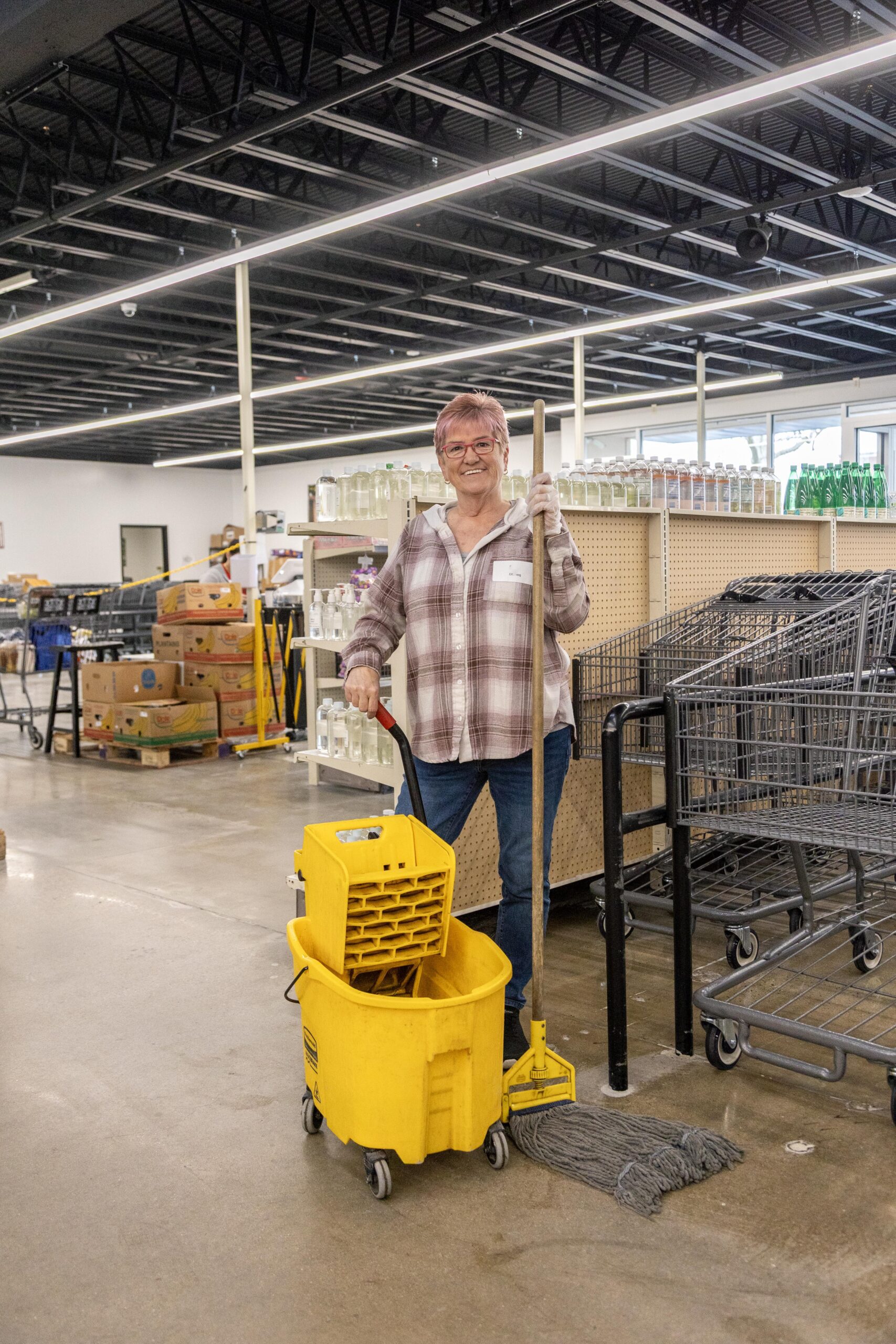 Senior woman smiling while holding a mop and standing next to a yellow mop bucket in a grocery store aisle, with shelves of products and shopping carts in the background.