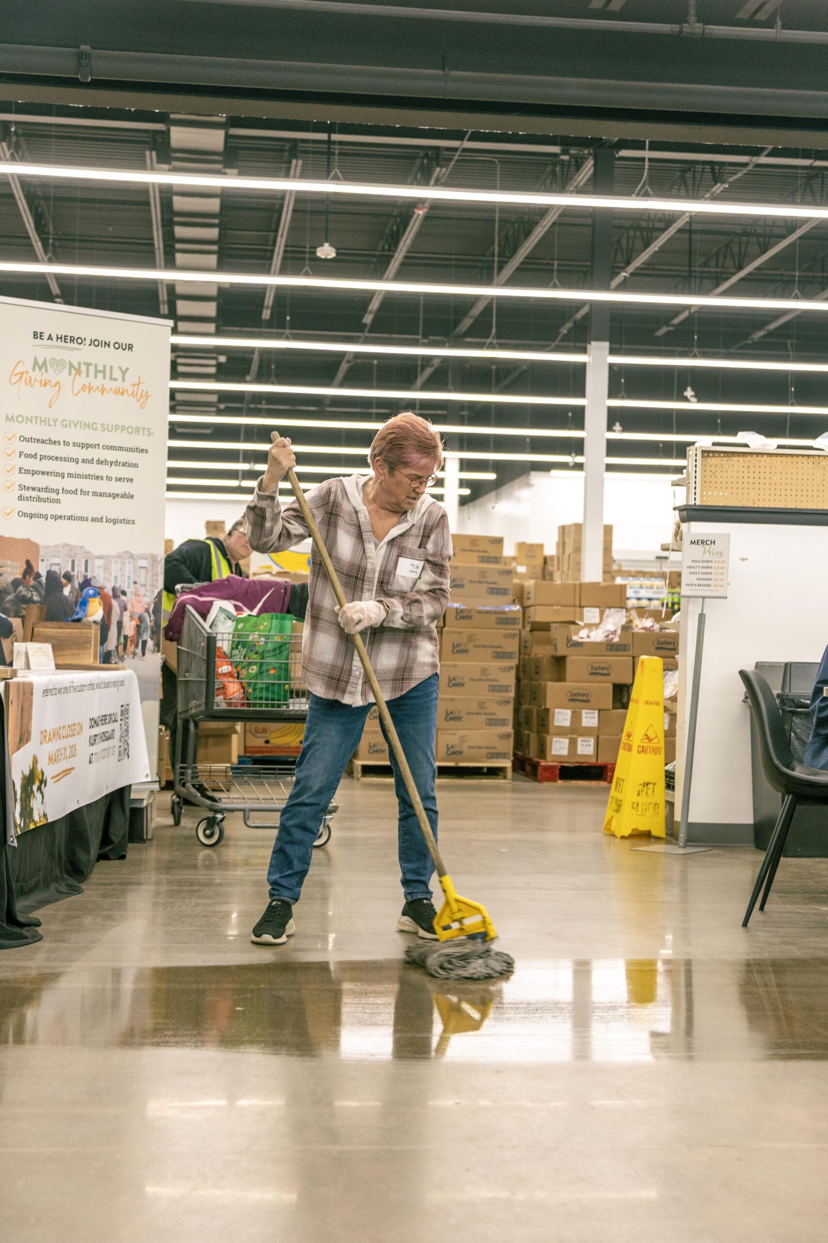 A volunteer mops the floor in a community center, surrounded by boxes and a display promoting monthly giving initiatives. The image captures the dedication to maintaining a clean and organized environment for community outreach programs.