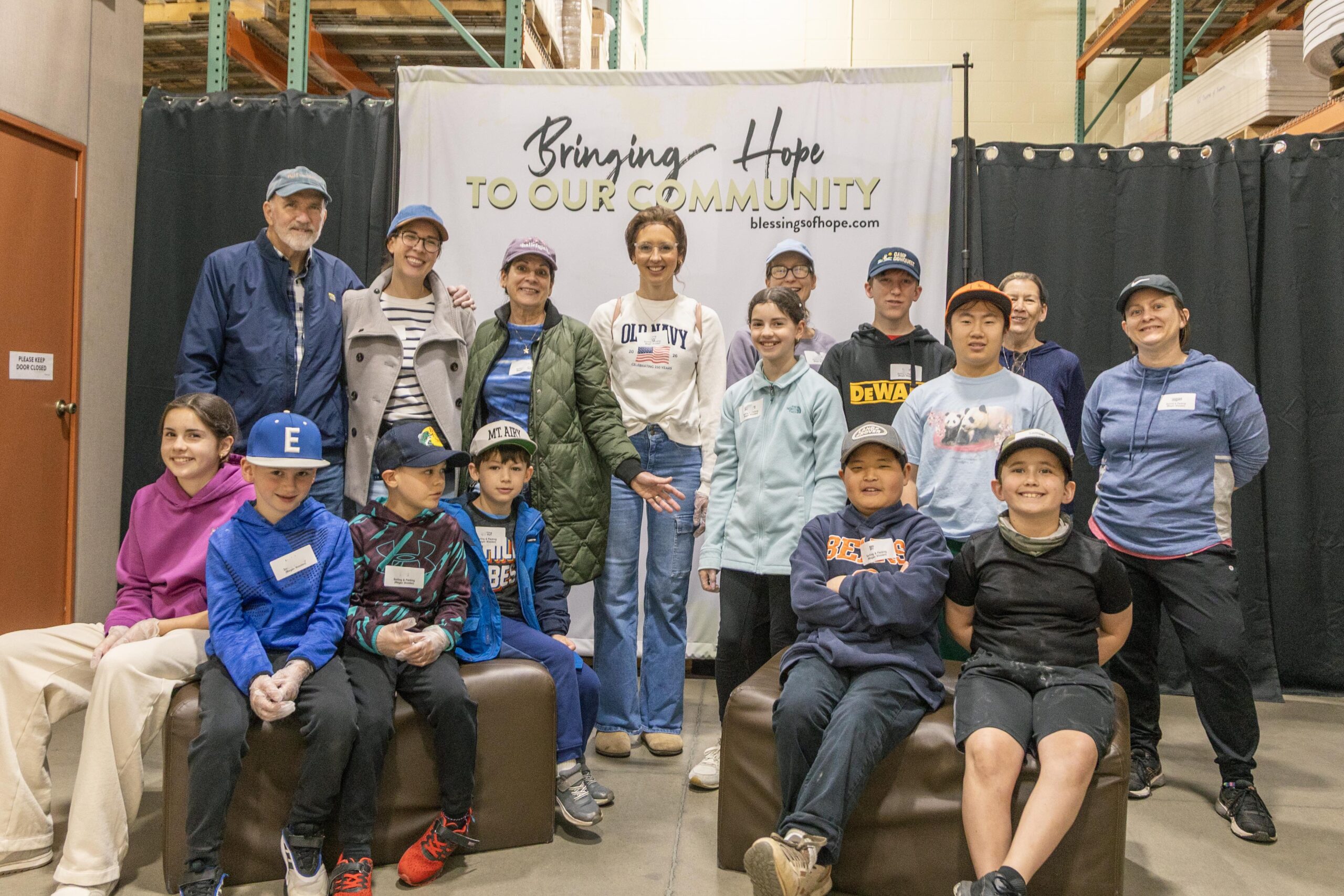 Group photo of volunteers and community members participating in a charitable event, standing and sitting together in front of a banner that reads "Bringing Hope to Our Community." The setting appears to be a warehouse or community center, with a focus on teamwork and community support.