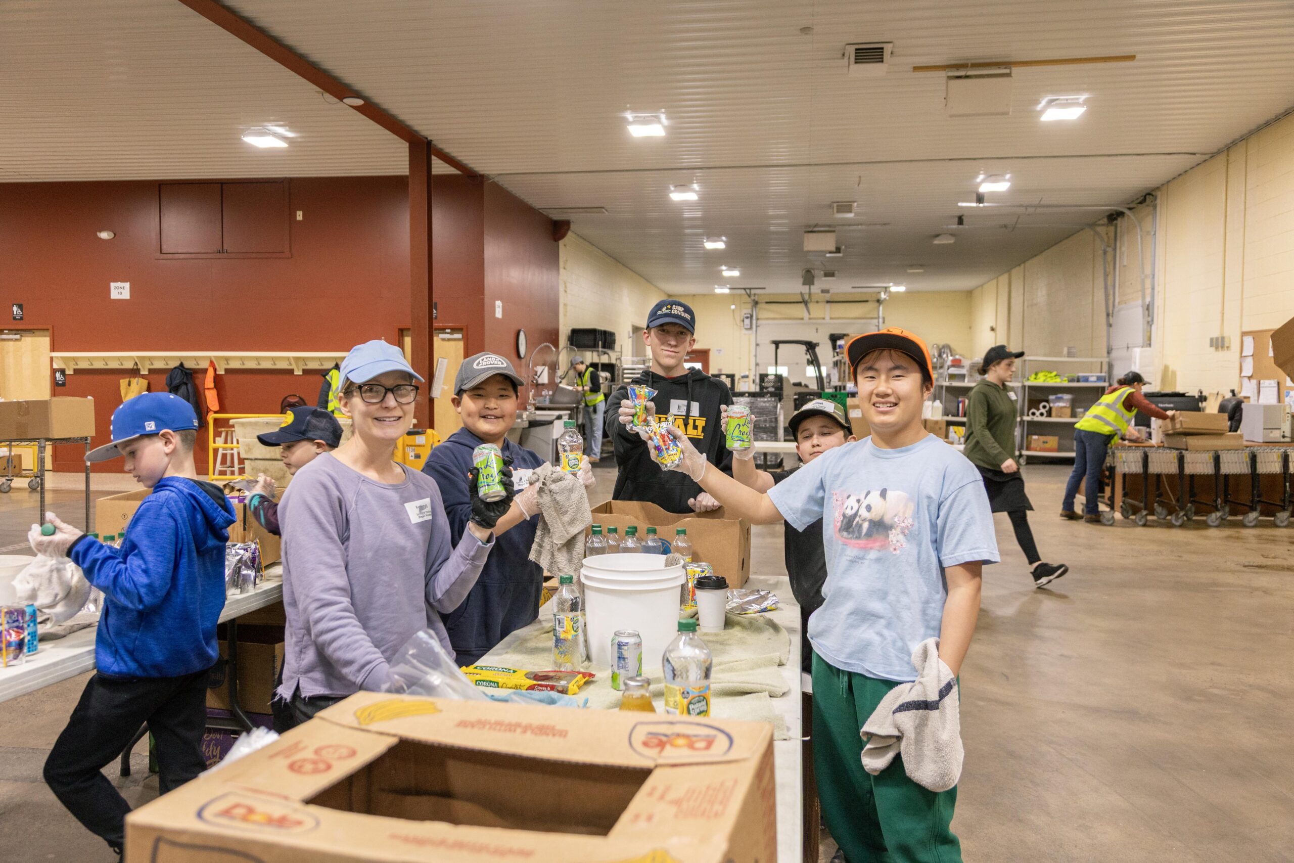 A group of children and a woman volunteer at a community recycling event, sorting and cleaning cans and bottles. They are smiling and holding up items they are working on, in a spacious indoor facility.