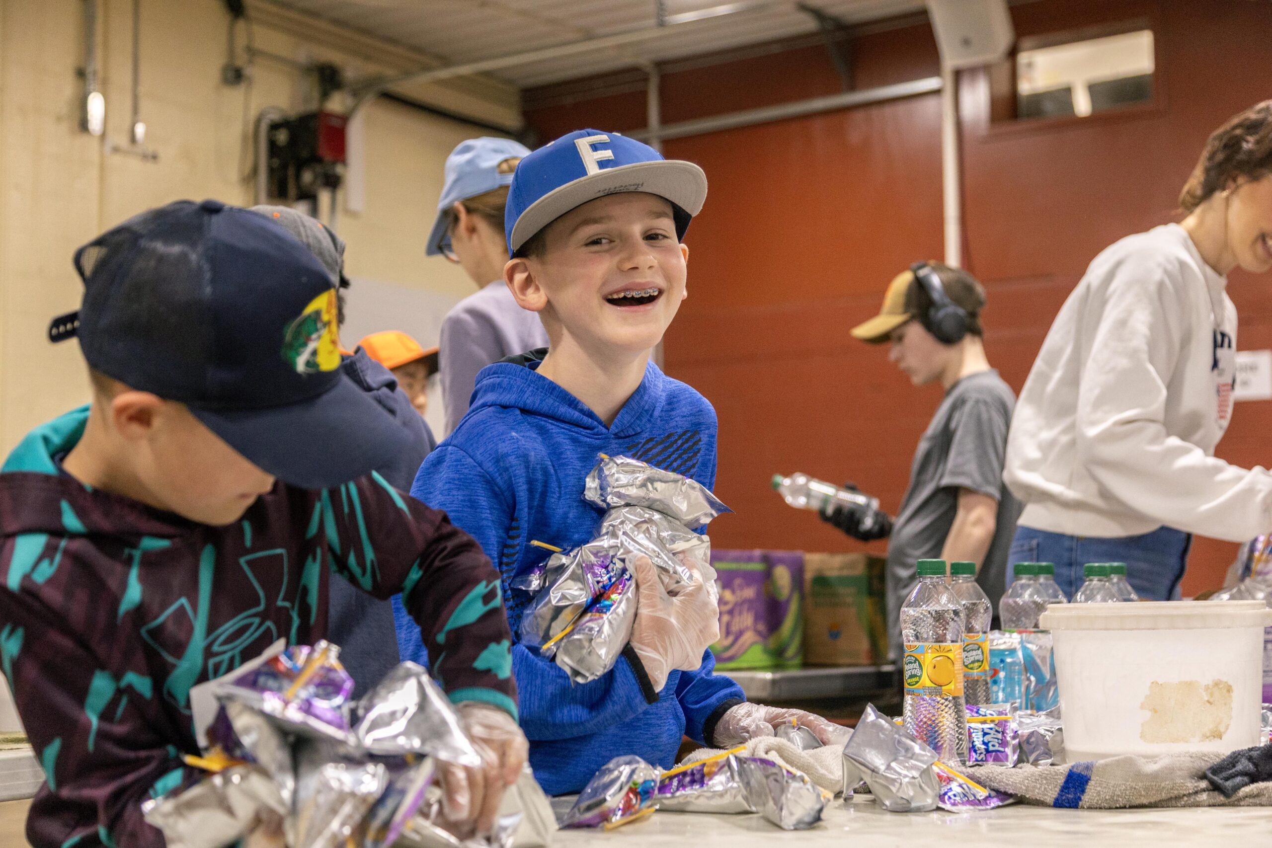 Children volunteering at a food packing event, smiling and working together to prepare snack packages, with colorful juice boxes and water bottles on the table.