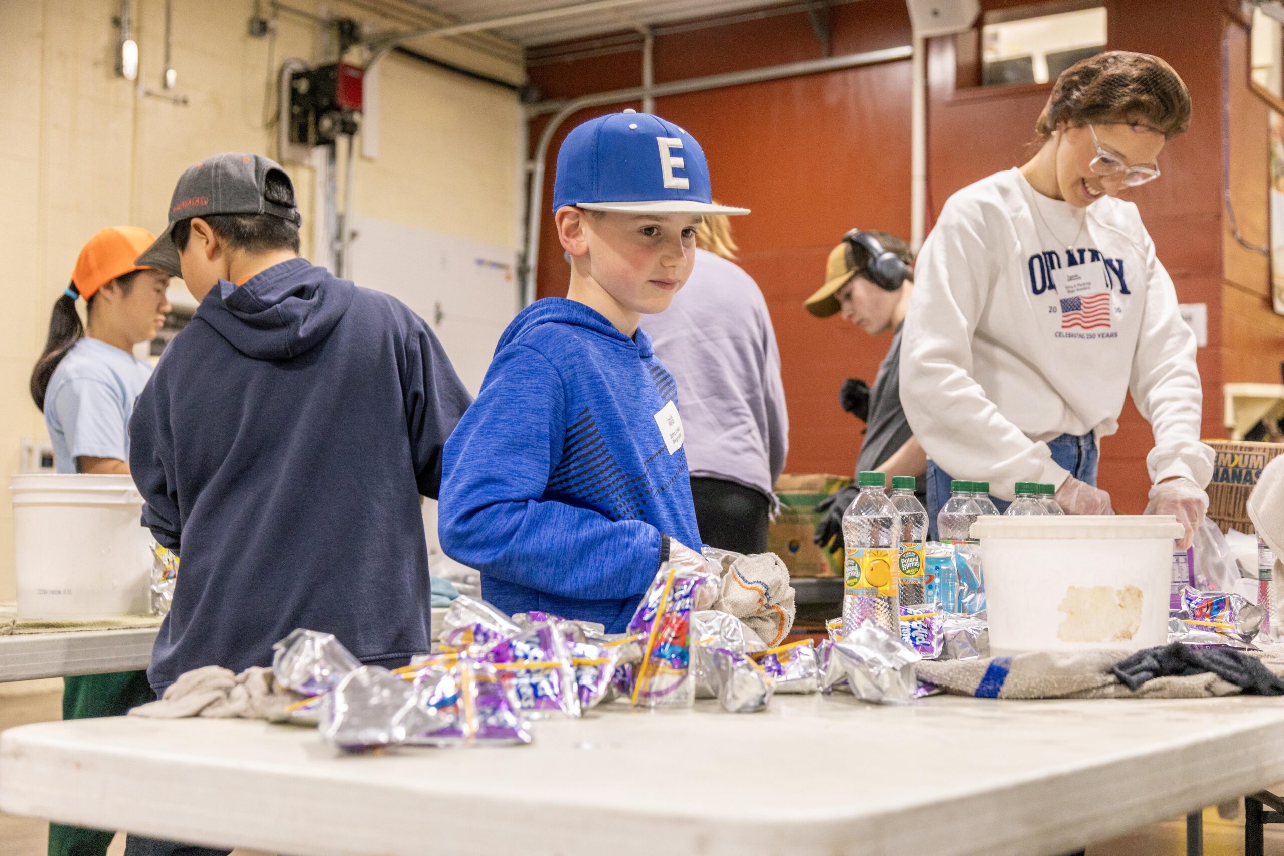 Children and volunteers participating in a community service event, preparing food items at a table with snacks and drinks in a spacious indoor setting.