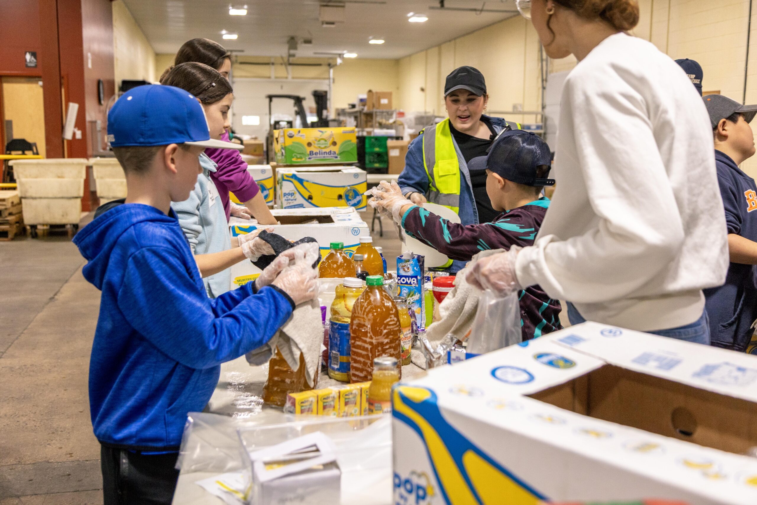 Children and adults volunteer at a food distribution center, packing beverages and food items into boxes while wearing gloves. The scene showcases teamwork and community involvement in supporting local families in need.