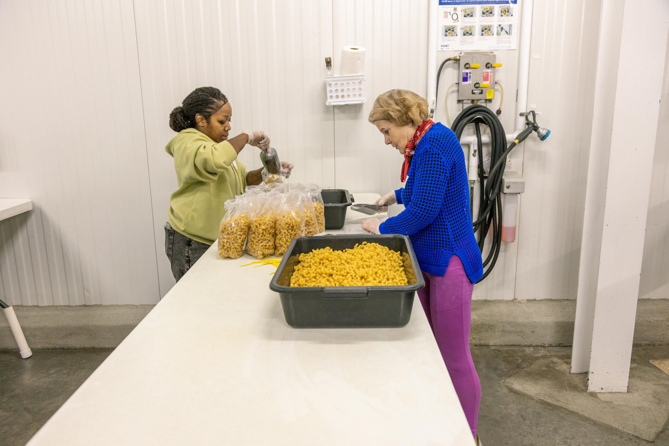 Two women working in a food processing facility, one scooping macaroni into bags while the other portions macaroni from a large tray. The setting features a clean, industrial kitchen environment with packaging supplies visible.