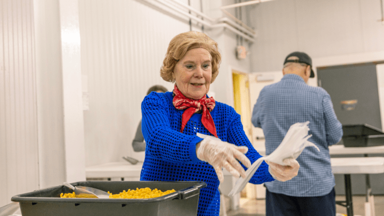 Senior woman in a blue sweater and red scarf serving macaroni and cheese in a community kitchen, wearing gloves and smiling, with volunteers in the background.