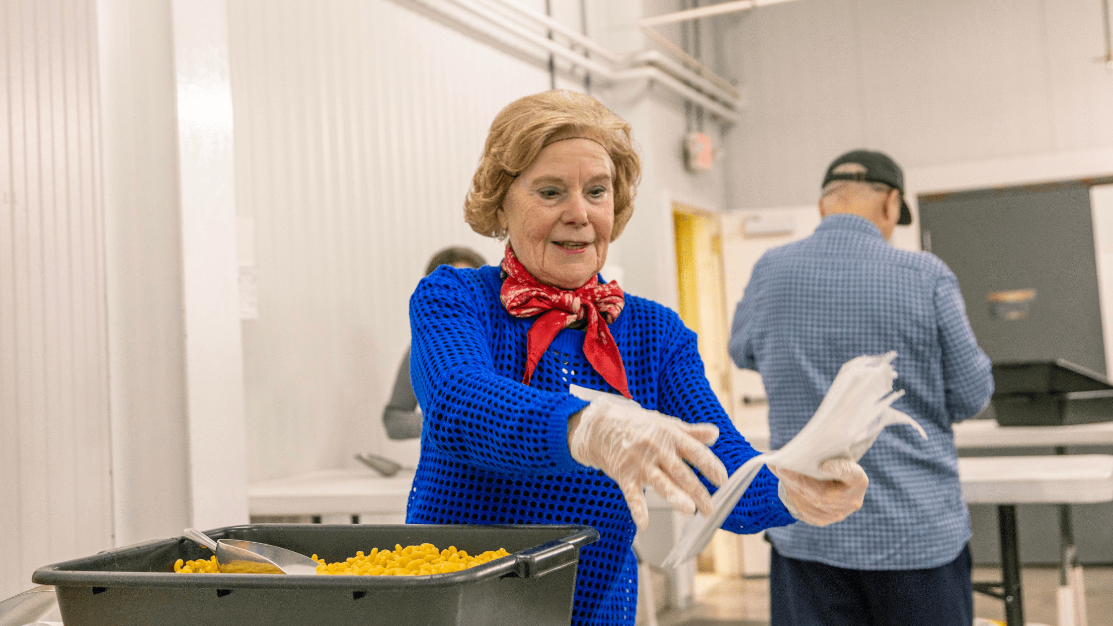 Senior woman in a blue sweater and red scarf serving macaroni and cheese in a community kitchen, wearing gloves and smiling, with volunteers in the background.