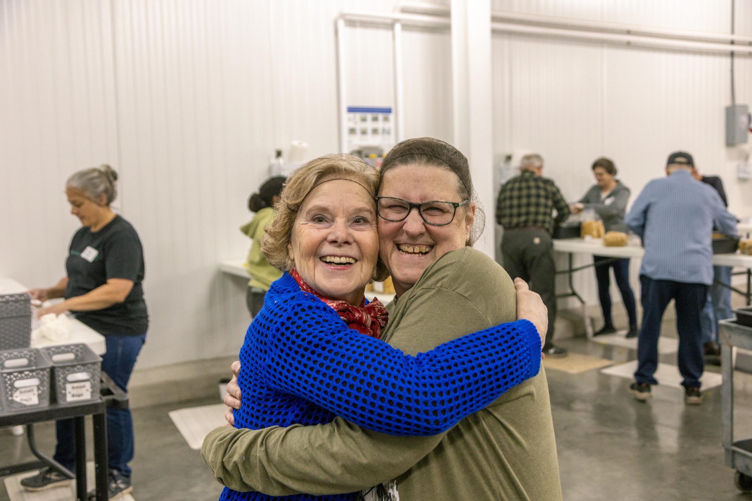 Two smiling women embrace joyfully in a community center, surrounded by volunteers engaged in food preparation activities. The setting reflects a warm, supportive atmosphere focused on community service and togetherness.