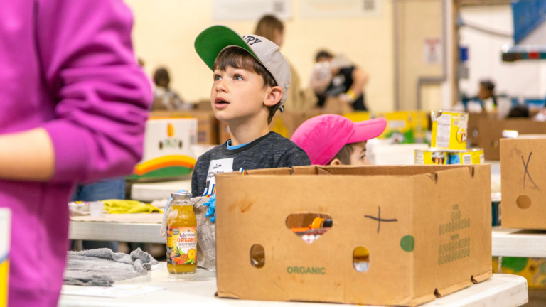 Young children participating in a community food distribution event, surrounded by cardboard boxes and organizing food items, with a focus on teamwork and learning about food security.