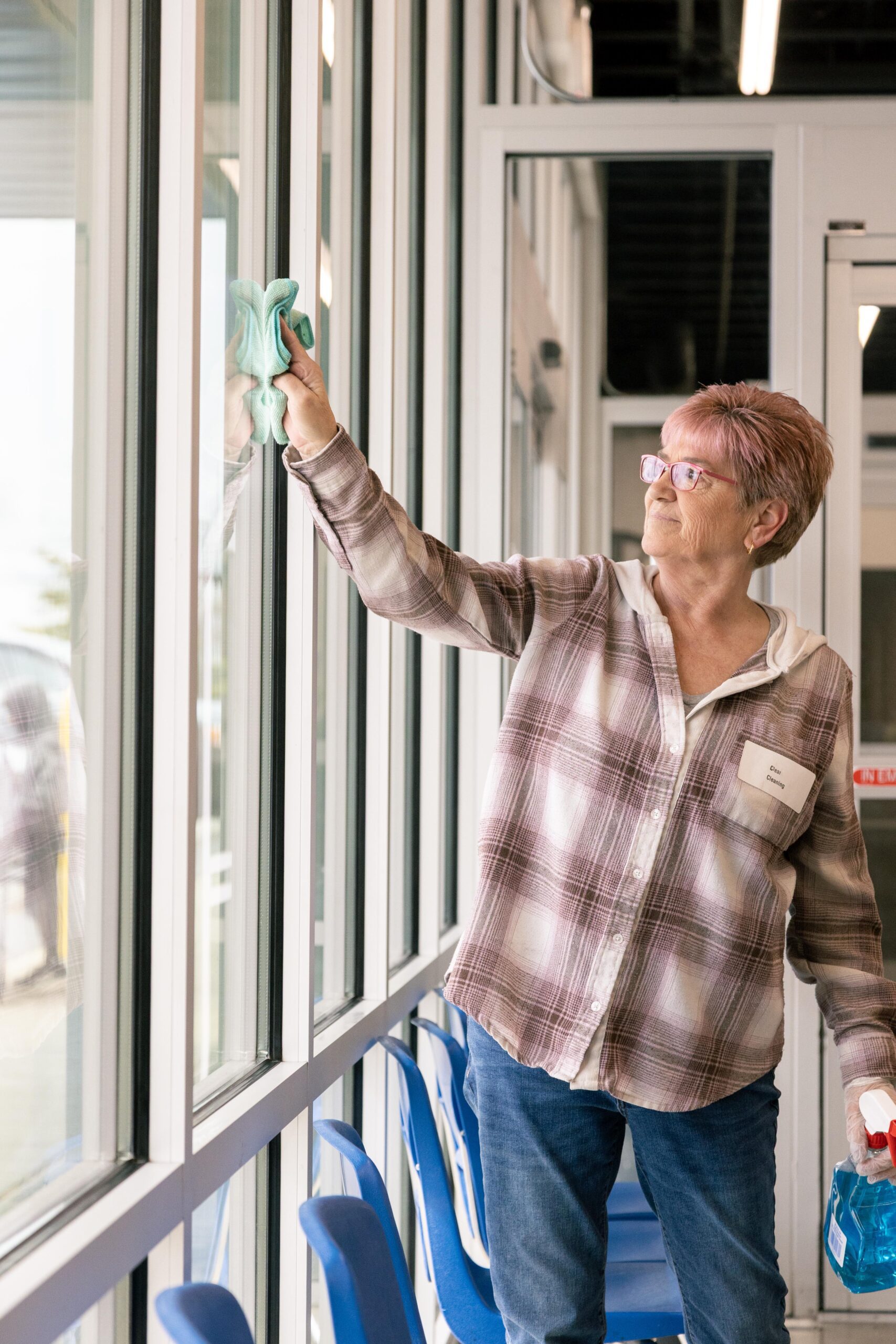 Senior woman cleaning large windows with a microfiber cloth, showcasing a bright, modern indoor space. She wears a plaid shirt and glasses, demonstrating a commitment to cleanliness and community service.