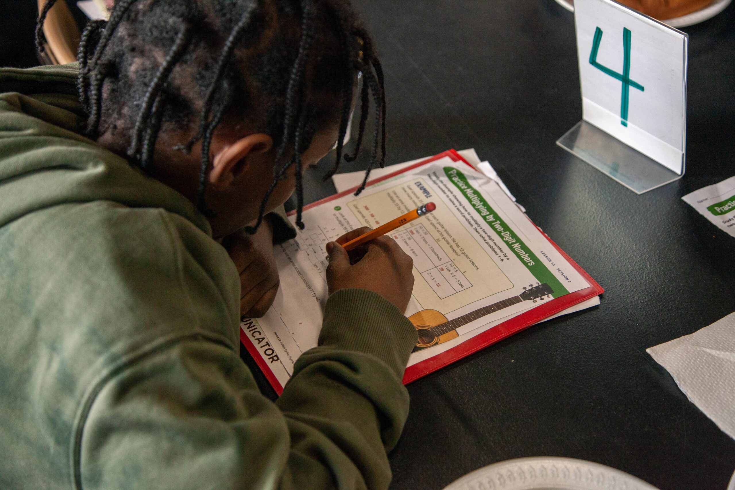 Child focused on completing math homework with a pencil on a worksheet featuring guitar illustrations, on a classroom table with a number sign displaying '4'.