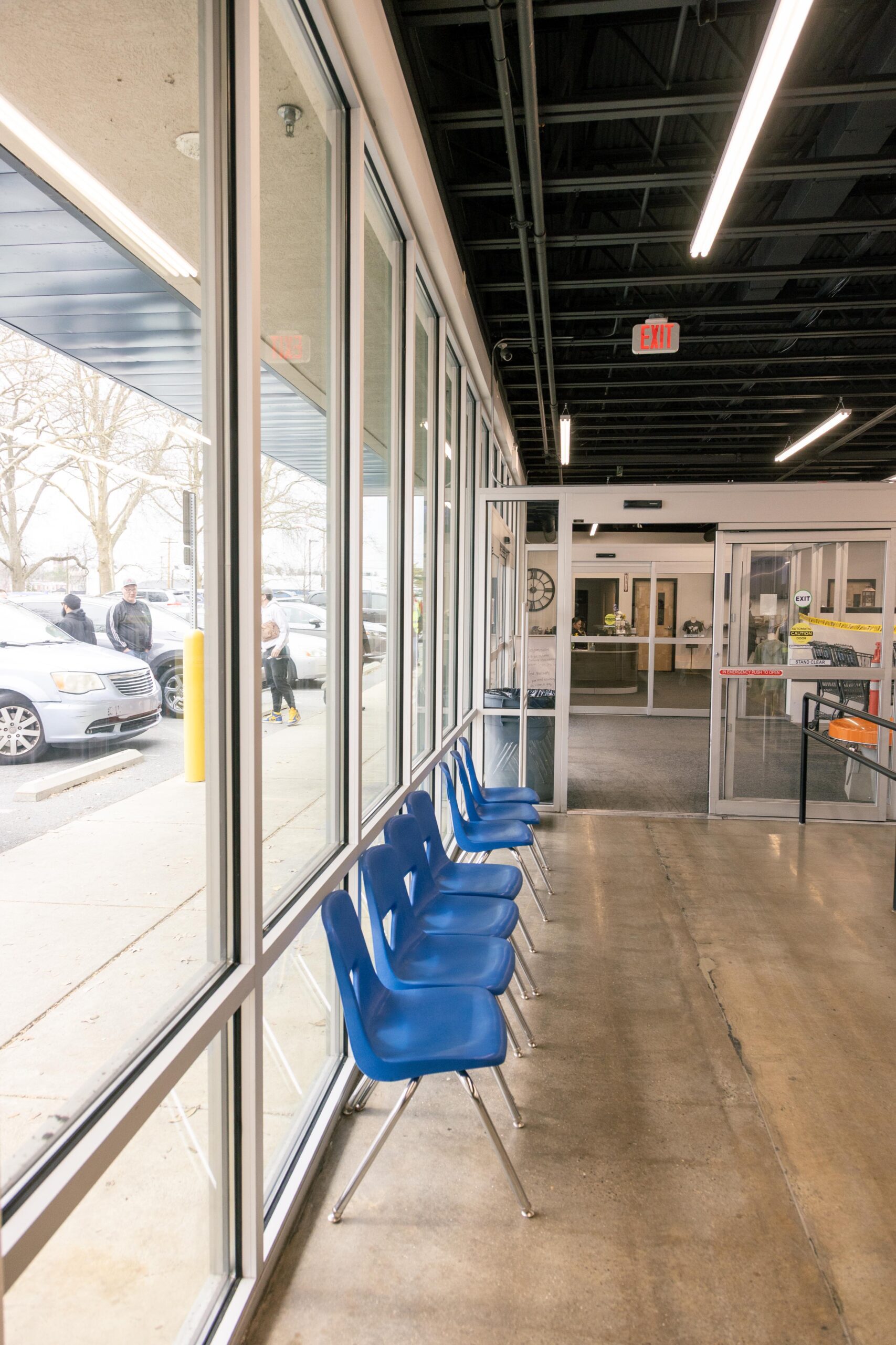 Interior view of a waiting area featuring blue chairs lined against large windows, allowing natural light. The setting includes a modern design with a concrete floor and an exit sign visible, creating a welcoming atmosphere for visitors.