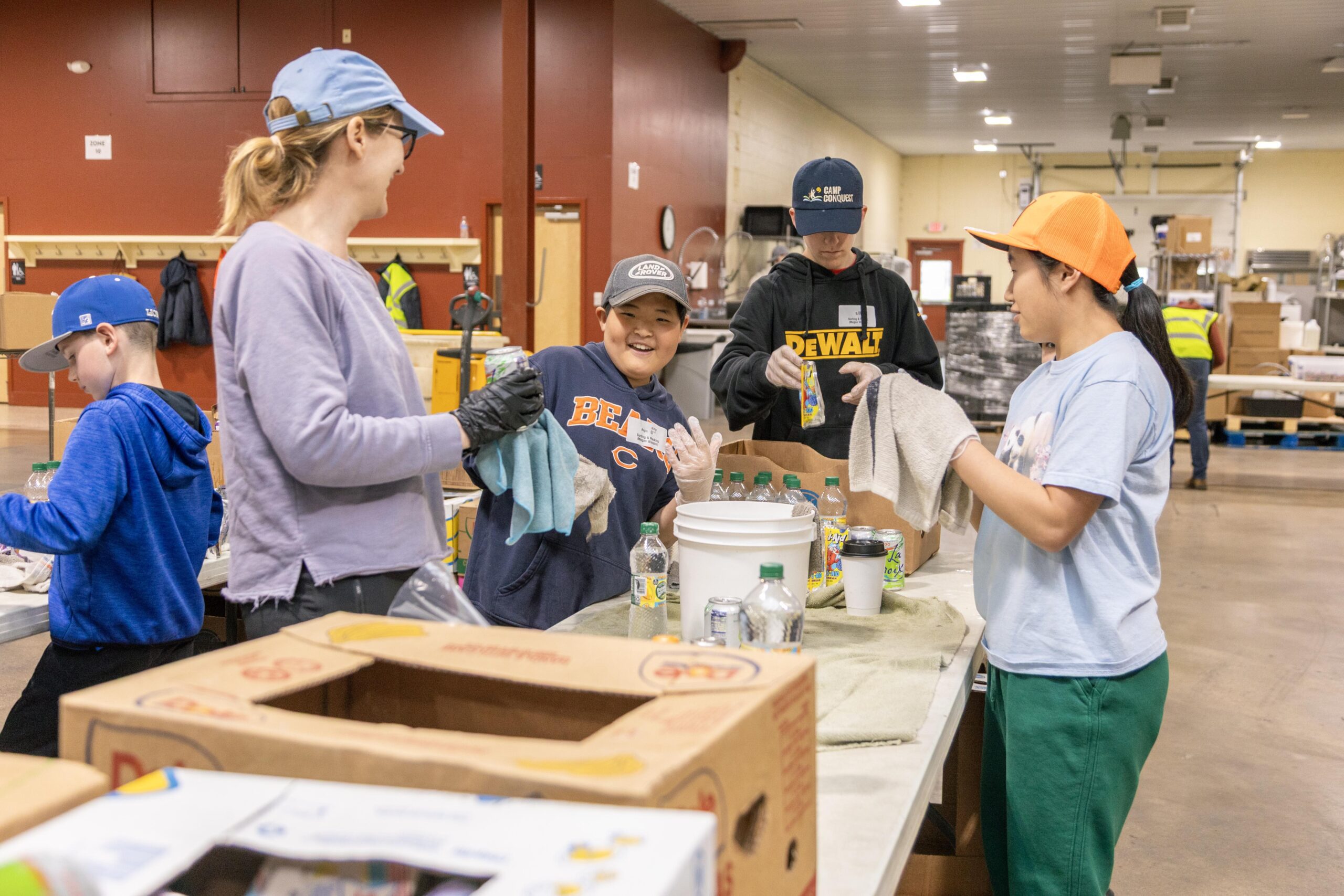 A group of volunteers, including children and adults, participate in a community service project, cleaning and sorting bottles and cans in a warehouse. The atmosphere is friendly and collaborative, showcasing teamwork in environmental efforts.