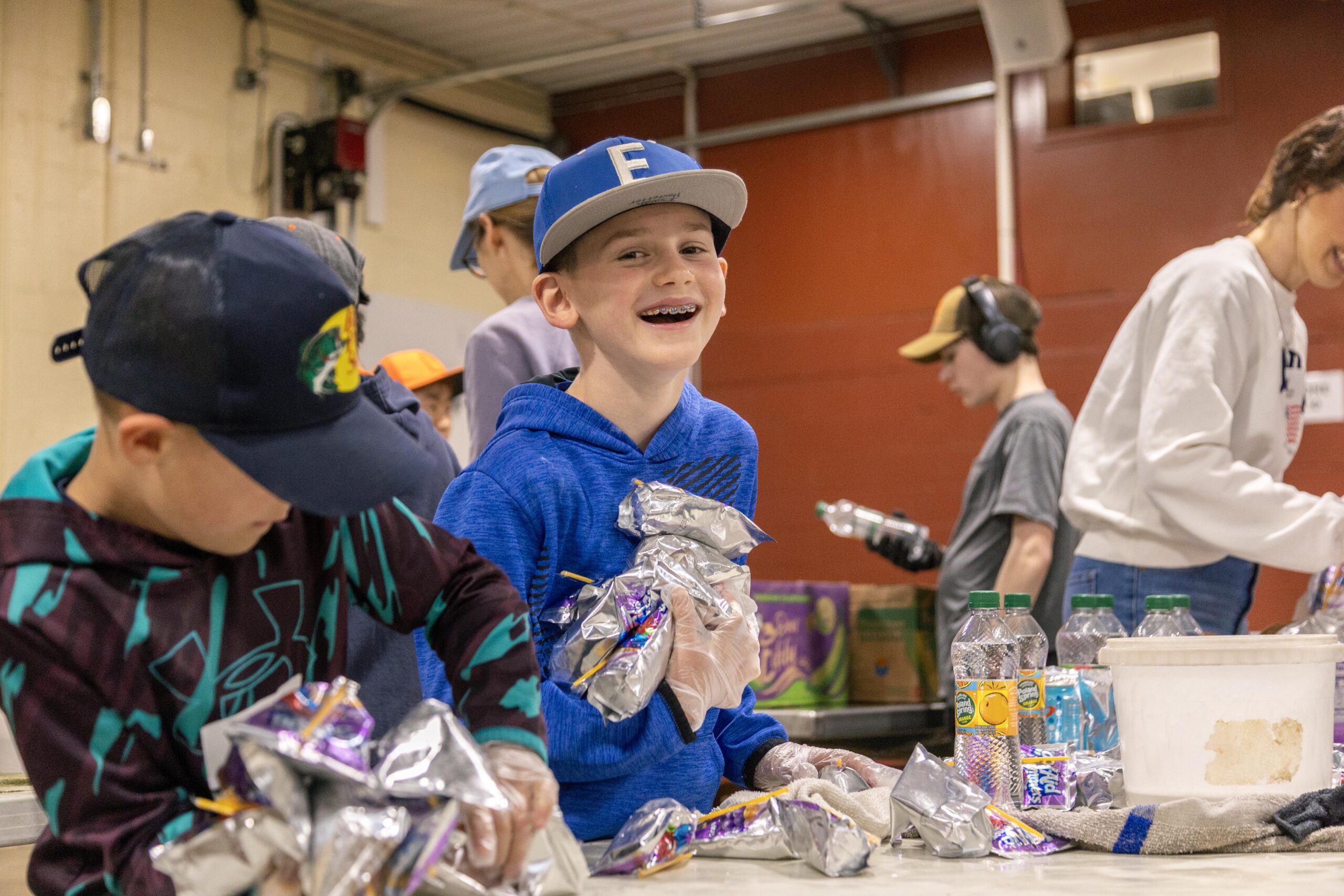 Children volunteering at a food packing event, smiling while holding wrapped snacks and working together in a community service project.