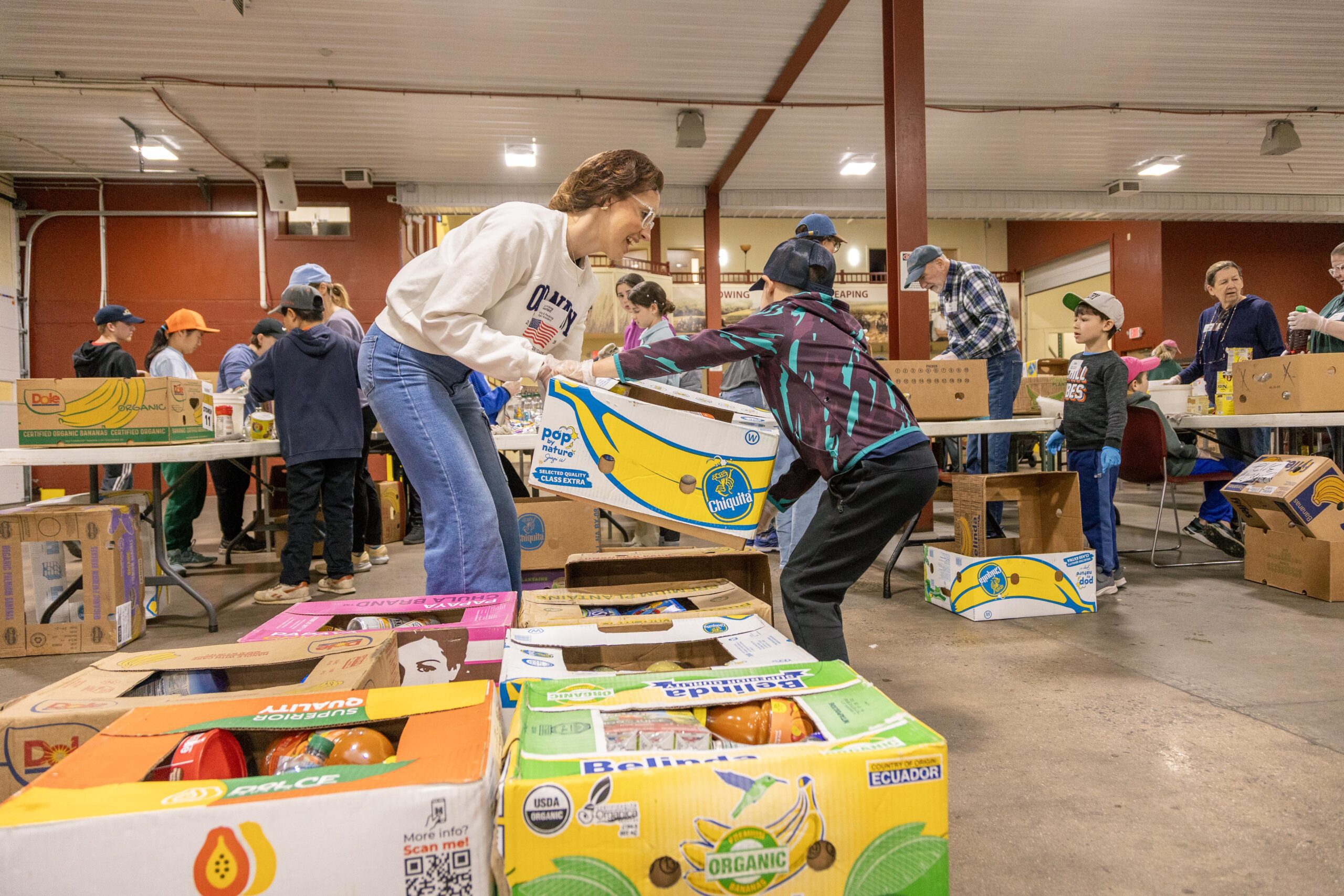 Volunteers sorting and packing food boxes at a community food drive event, with children and adults working together in a spacious indoor setting.