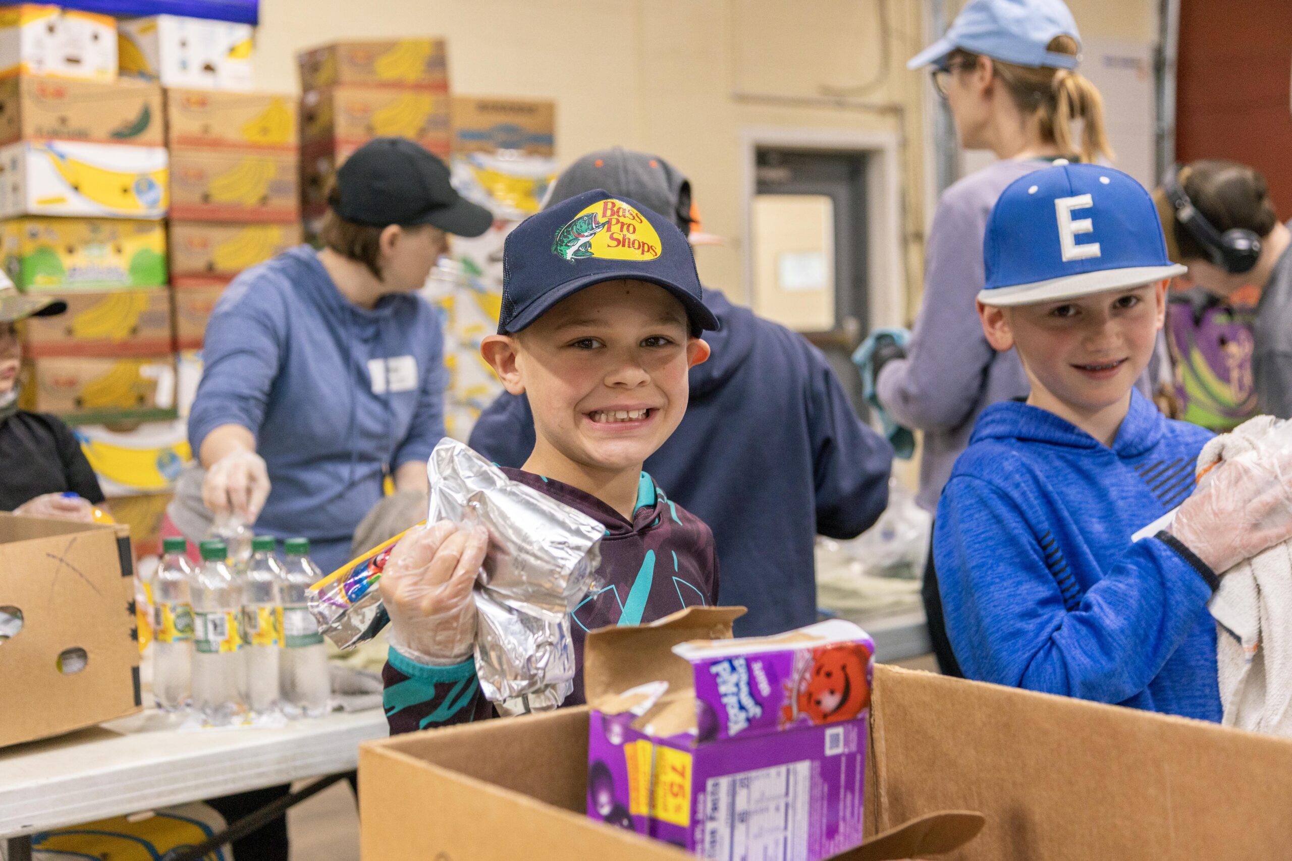 Children volunteering at a food distribution event, smiling while helping pack boxes with snacks and beverages. In the background, other volunteers are organizing food items in a community service setting.