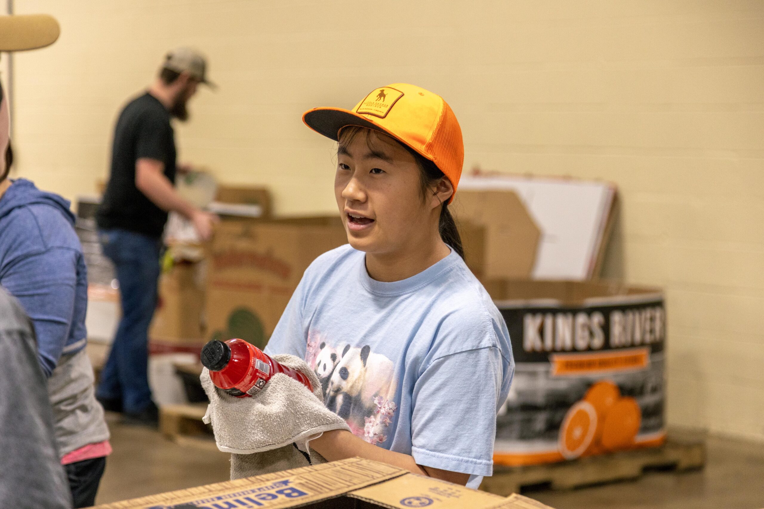 A young volunteer in an orange cap holds a bottle and towel while engaging in a community service event, with cardboard boxes and other volunteers in the background.