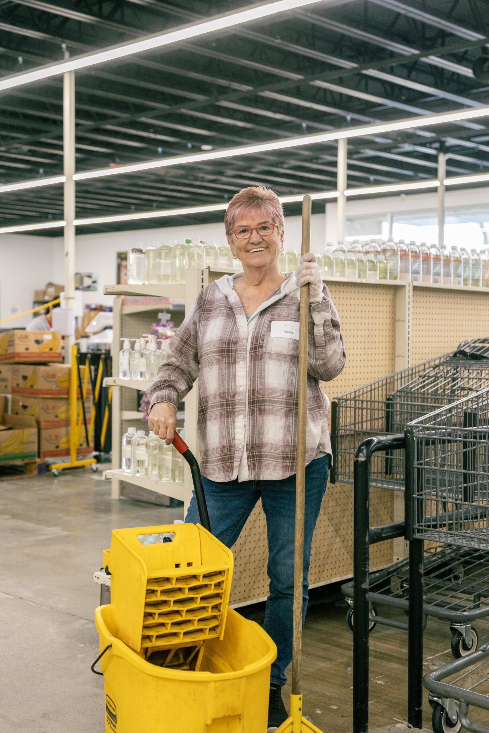Smiling older woman holding a mop and standing next to a yellow mop bucket in a retail store aisle, surrounded by shelves stocked with bottles and shopping carts.