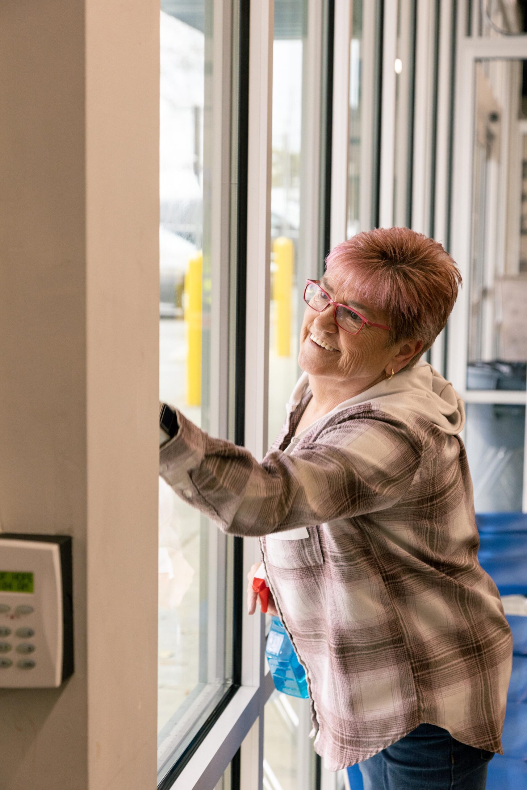 Senior woman with short pink hair and glasses smiles while cleaning a large window in a bright indoor space. She wears a plaid shirt and appears cheerful and engaged in her task.