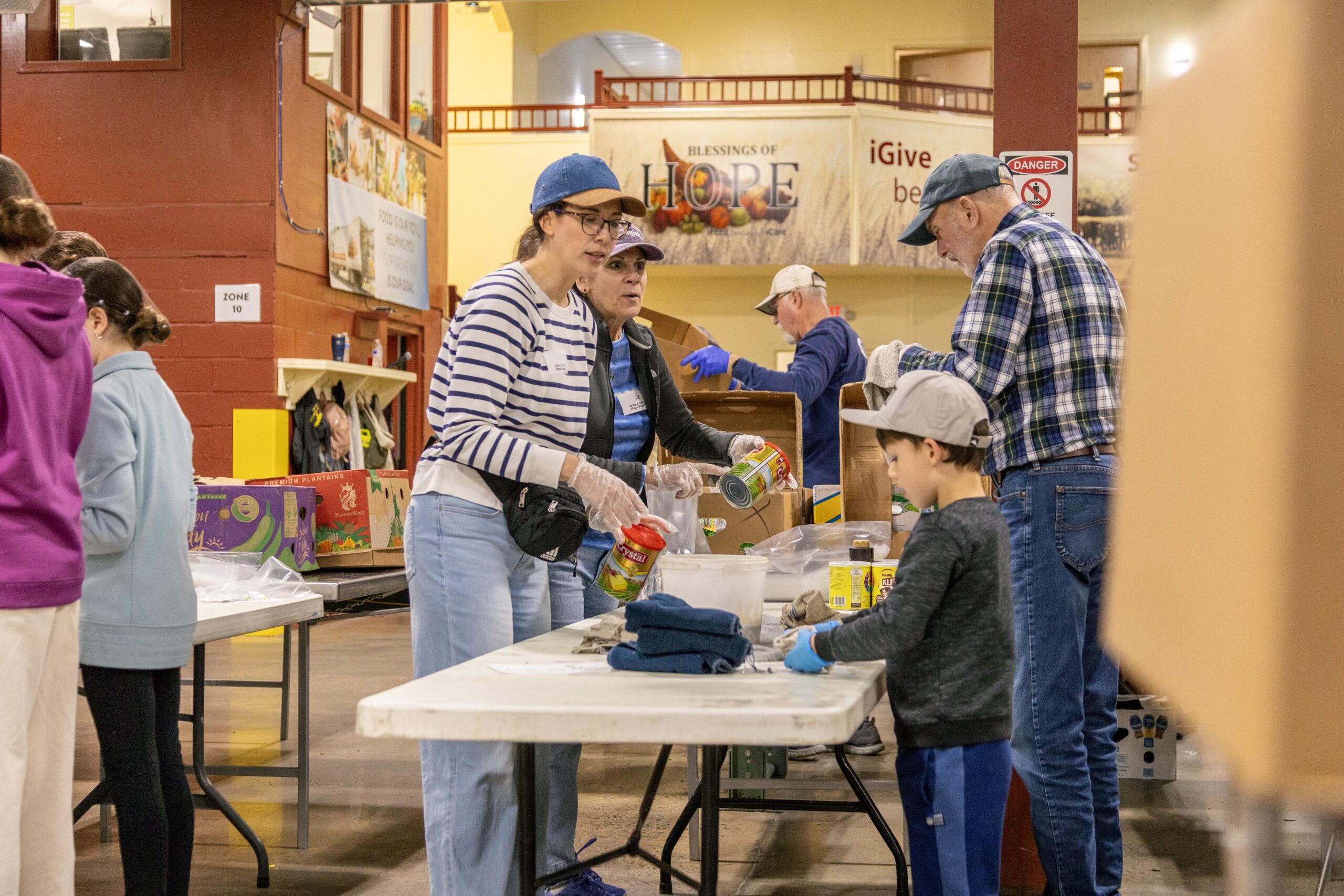 Volunteers of various ages participate in a food distribution event, sorting and organizing canned goods and other food items at a community center. The environment is busy and collaborative, highlighting the spirit of community service.