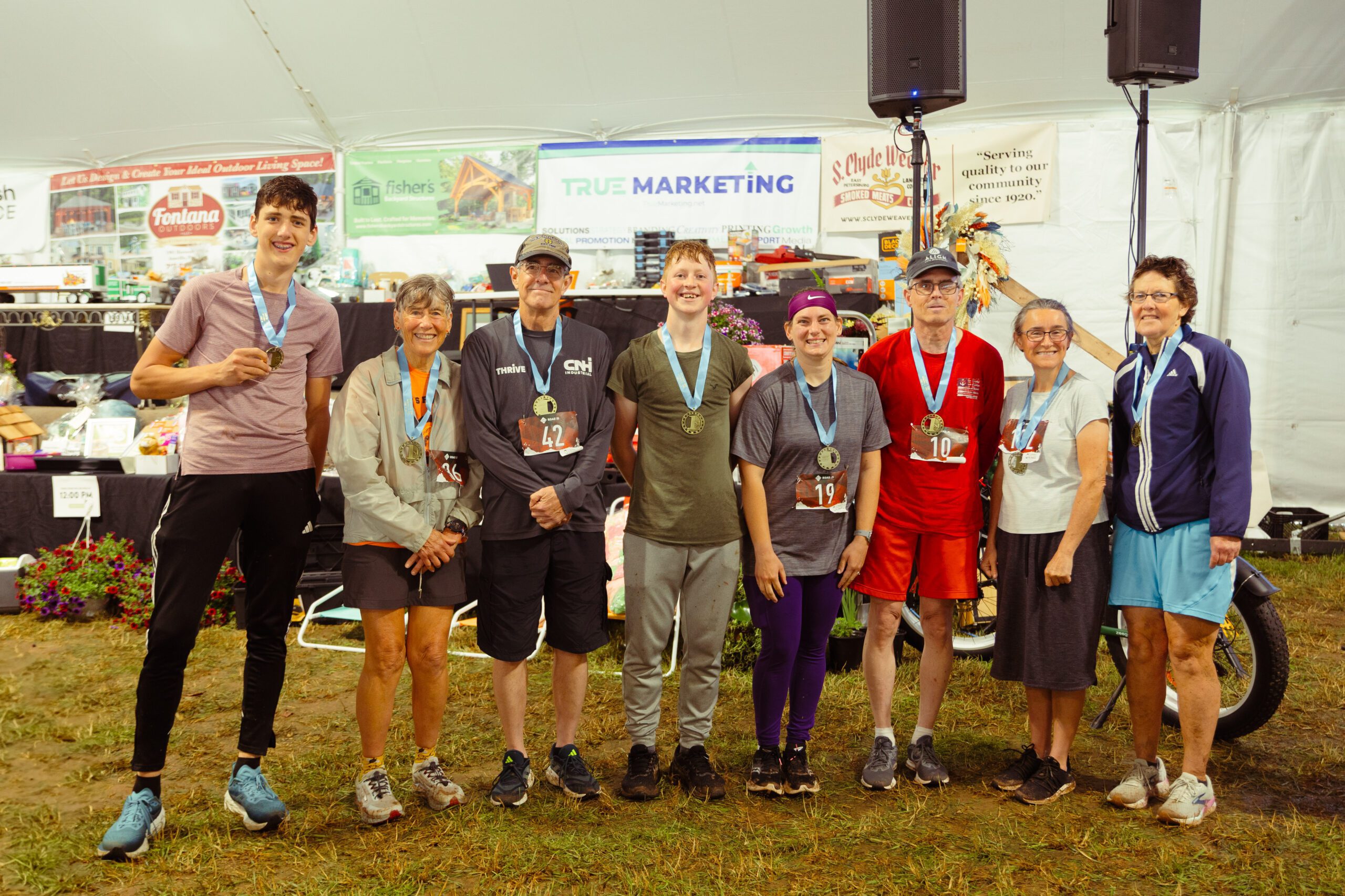 Group of diverse athletes celebrating after a race, wearing medals and race bibs in a tent decorated with sponsor banners. The group includes men and women of various ages, showcasing a community spirit and achievement in sports.