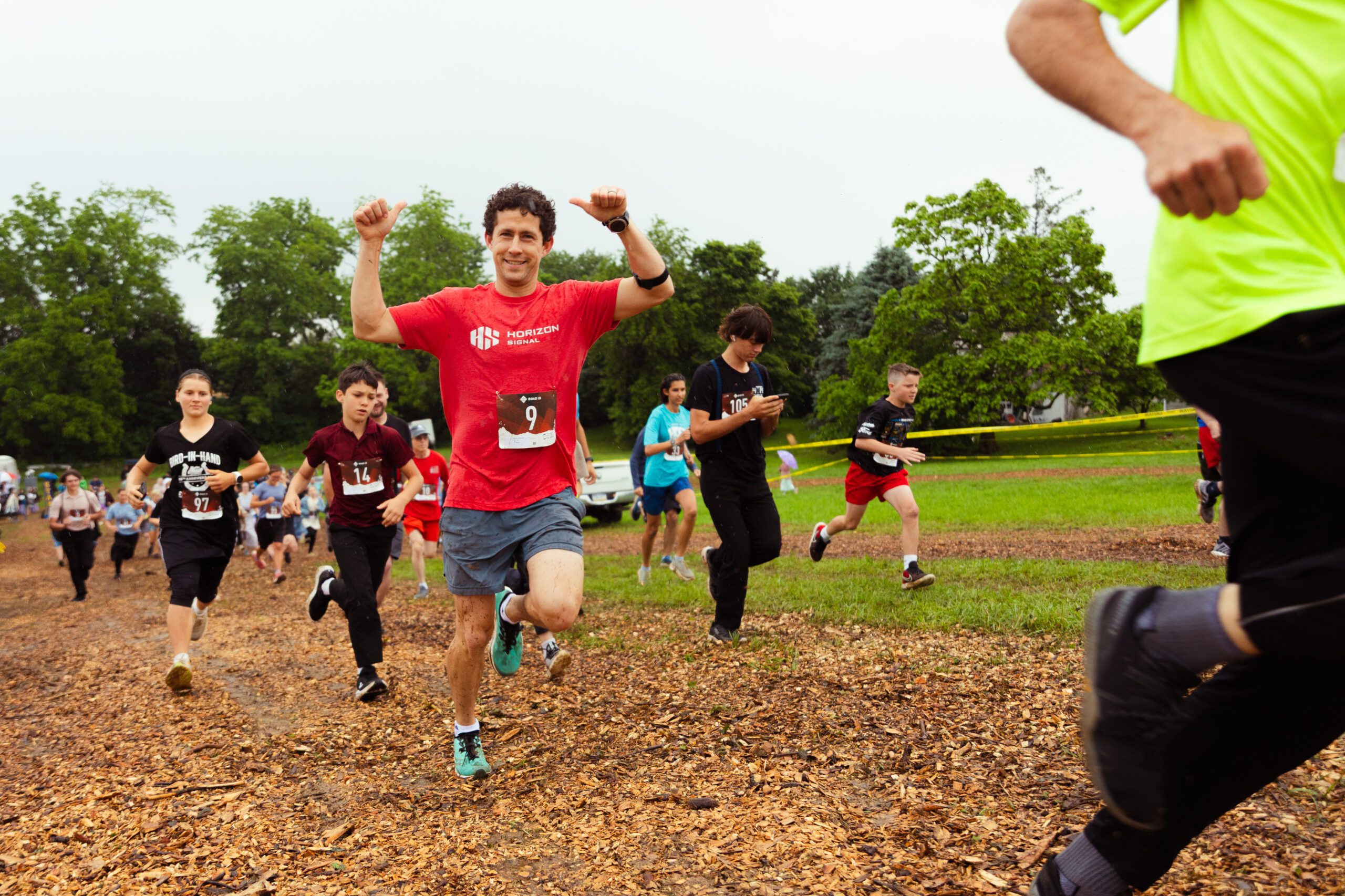 Participants in a fun run event sprinting on a wooded trail, showcasing enthusiasm and teamwork. The scene features diverse runners wearing numbered bibs and athletic attire, with a vibrant green landscape in the background.
